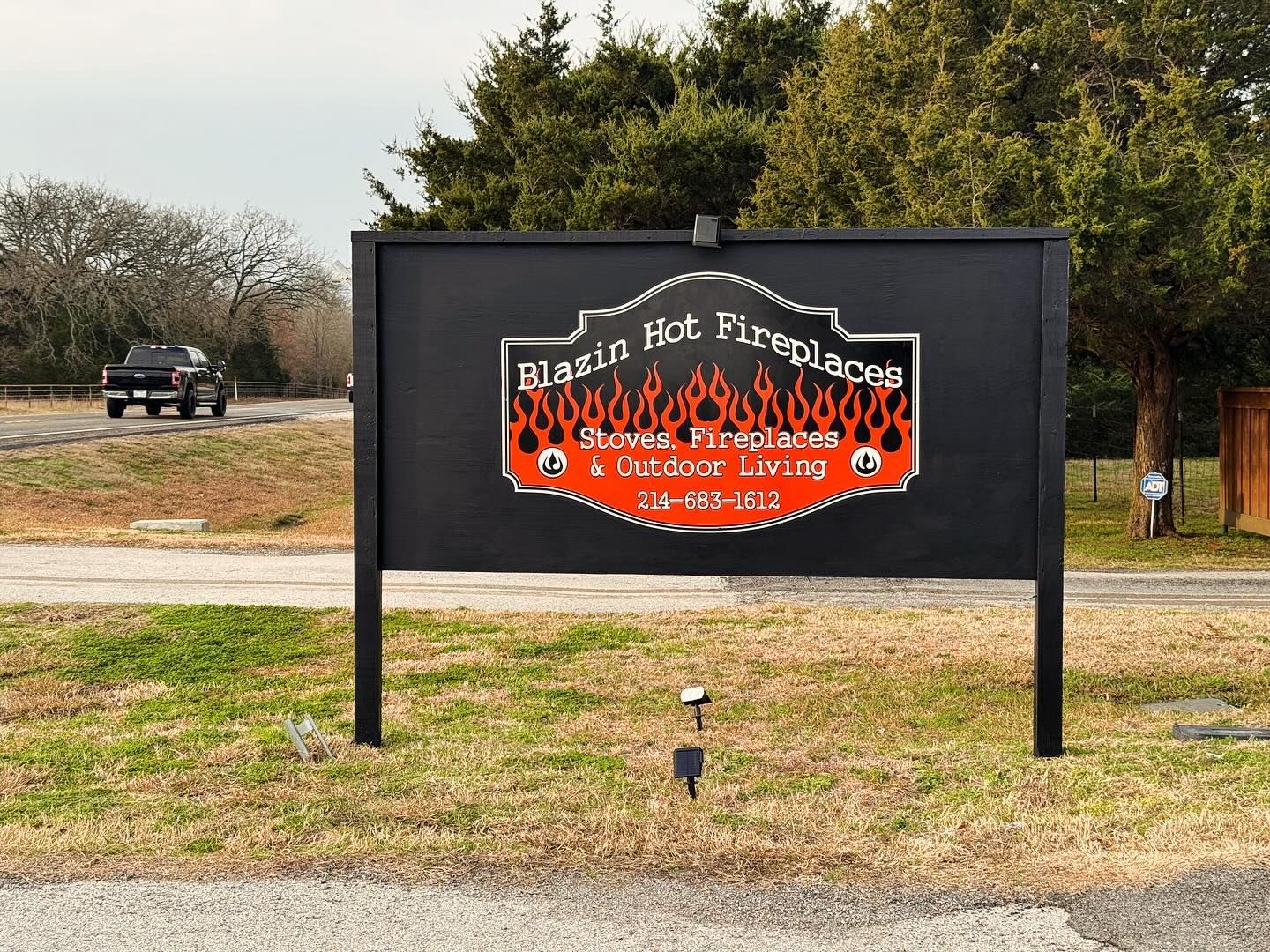 Black Hot Firewood sign with red flame graphic on a roadside in a grassy area