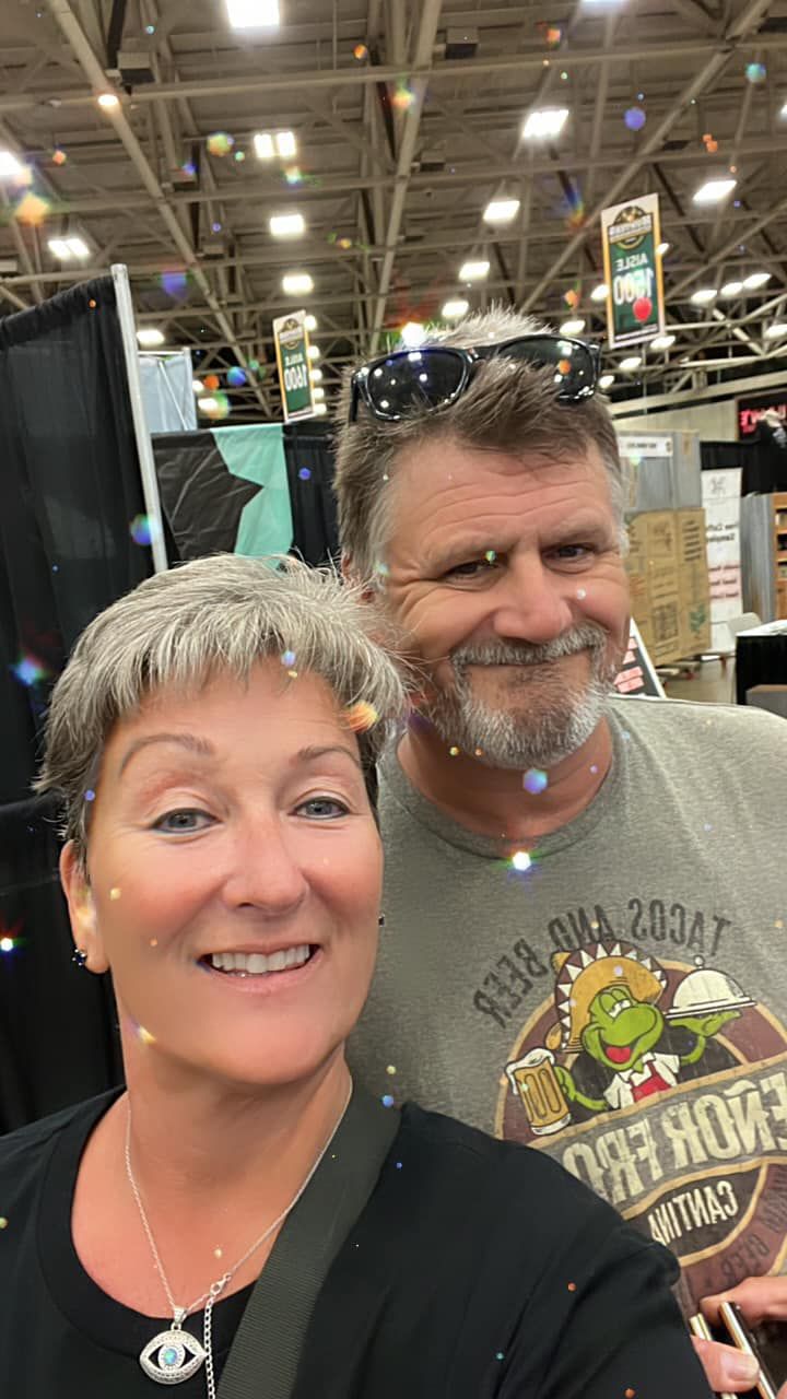 Two smiling people taking a selfie at an indoor expo booth, with bright overhead lights and display banners.