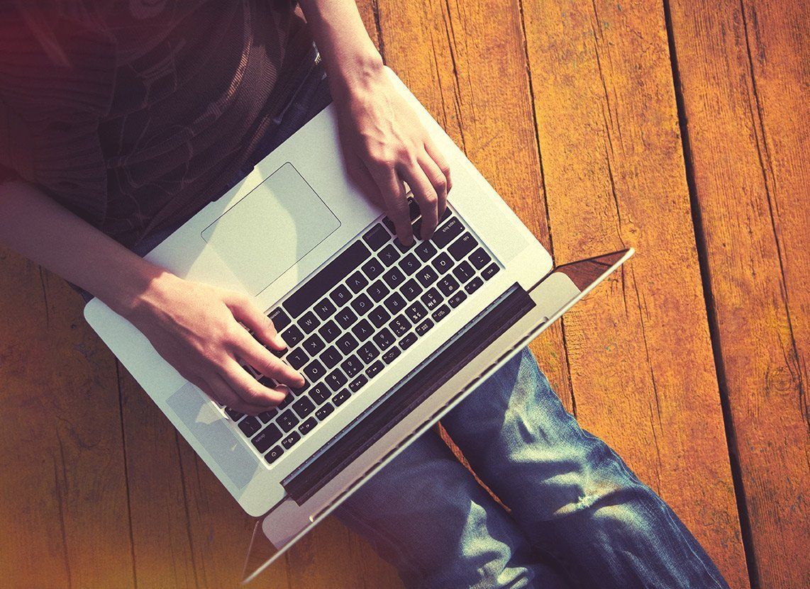 Hands typing on a laptop on a wooden floor, viewed from above.