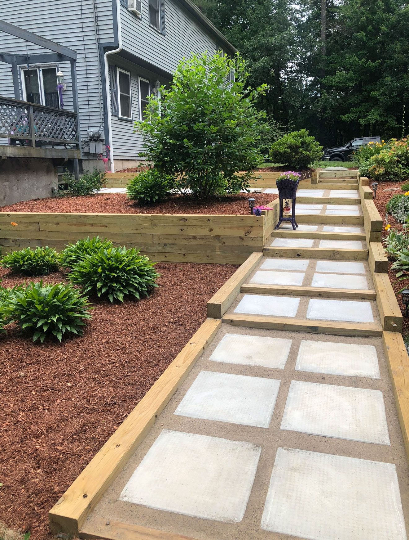 A path of square concrete pavers with wooden borders leads up through a mulched landscape to a grey house.