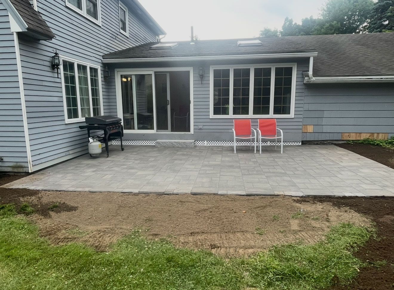 A gray house with a newly installed gray paver patio, a black grill, and two red chairs against the back wall.
