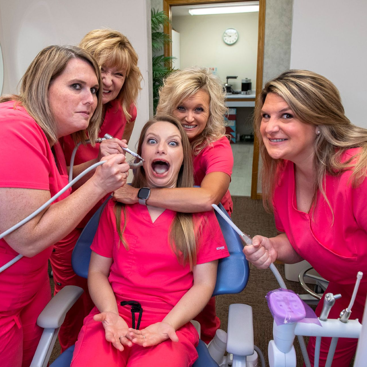 A group of women in pink scrubs are examining a woman 's teeth in a dental chair.