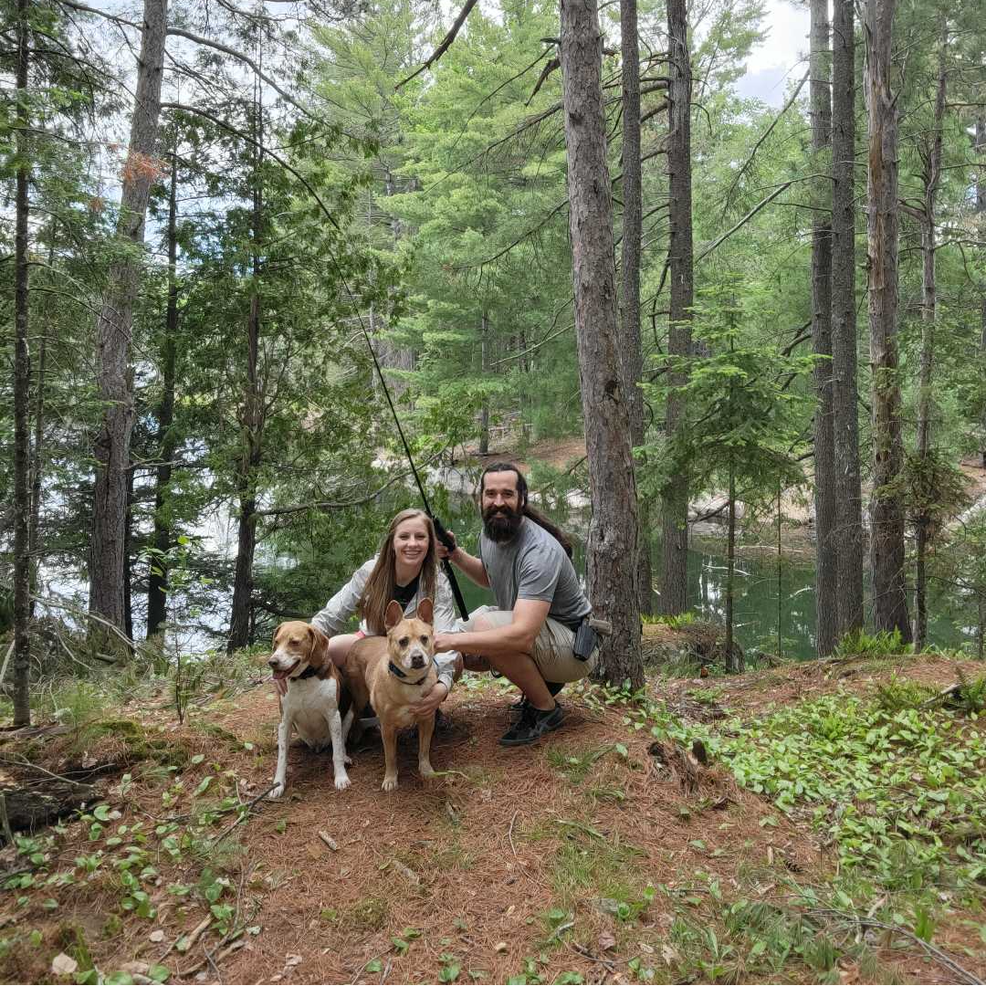 A man and a woman are posing for a picture with their dogs in the woods.