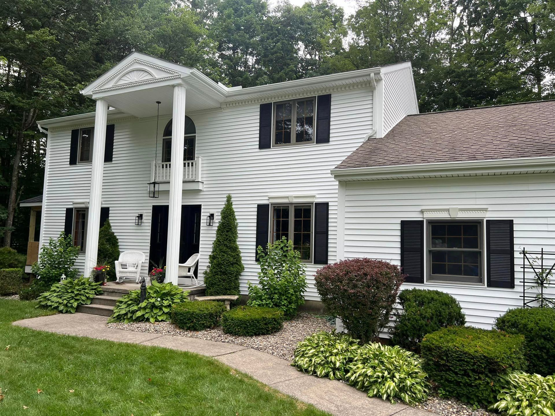 A large white house with black shutters is surrounded by trees and bushes