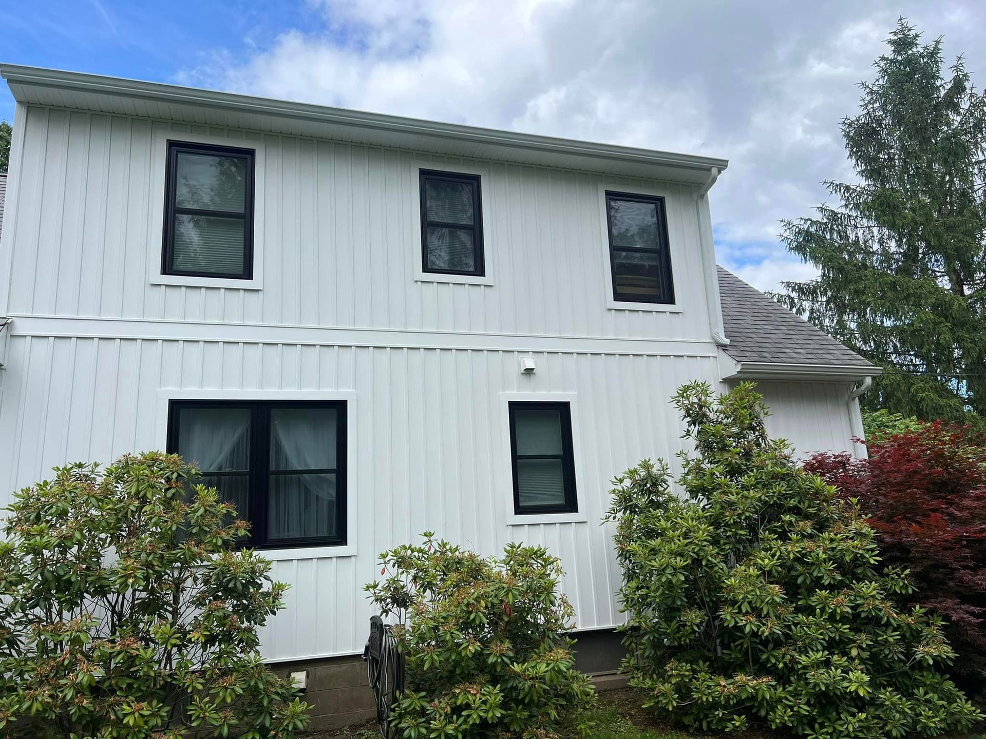A white house with black windows is surrounded by bushes and trees