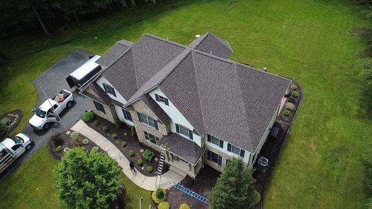 Aerial view of a two-story house with a brown roof. Two white trucks are parked on a driveway.