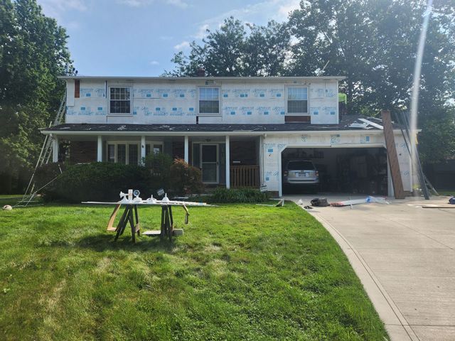 House exterior under renovation: blue wrap, exposed roof, garage, tools, green grass, sunny day.