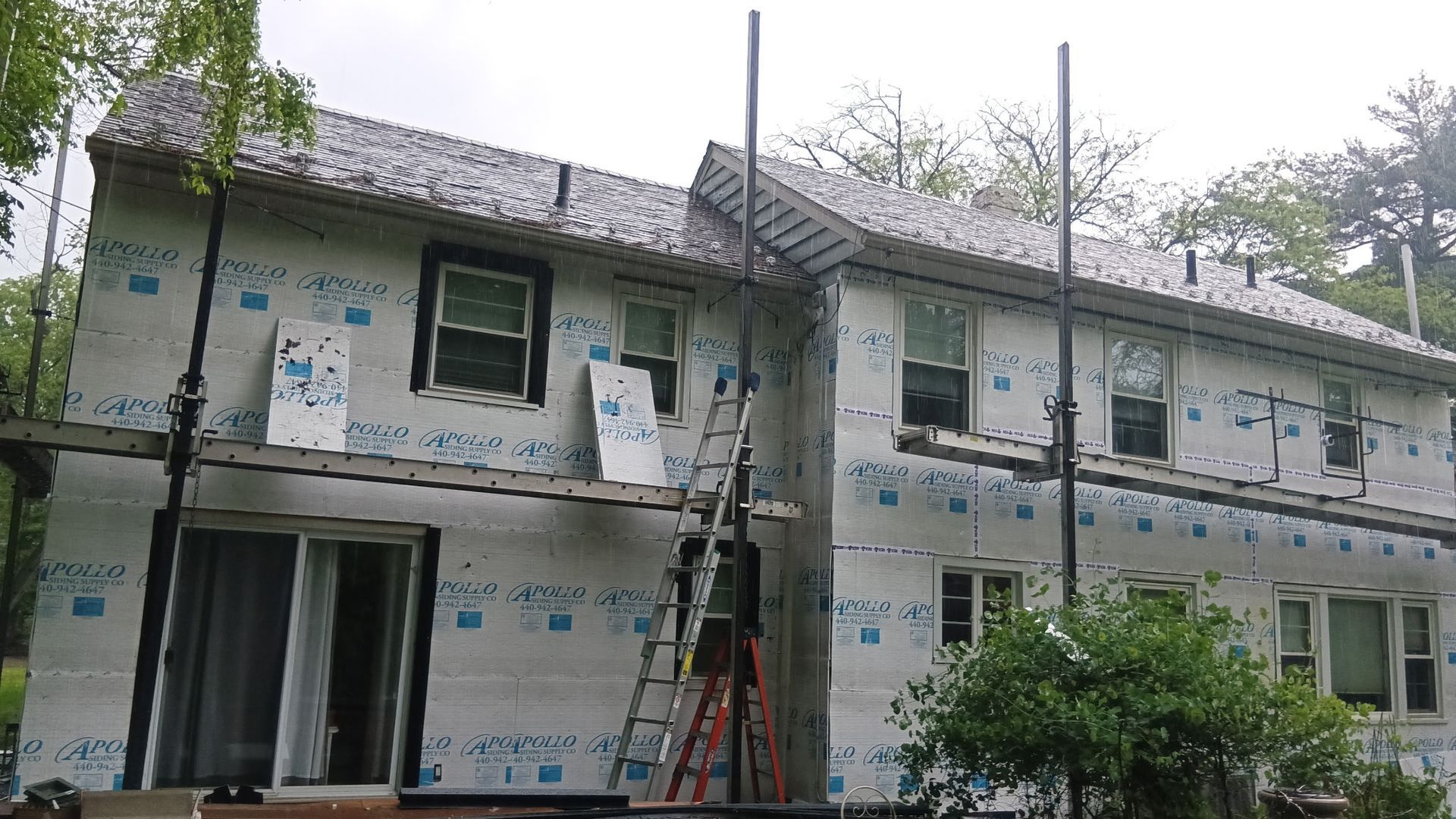 House exterior under construction, with scaffolding and insulation wrap; cloudy sky.