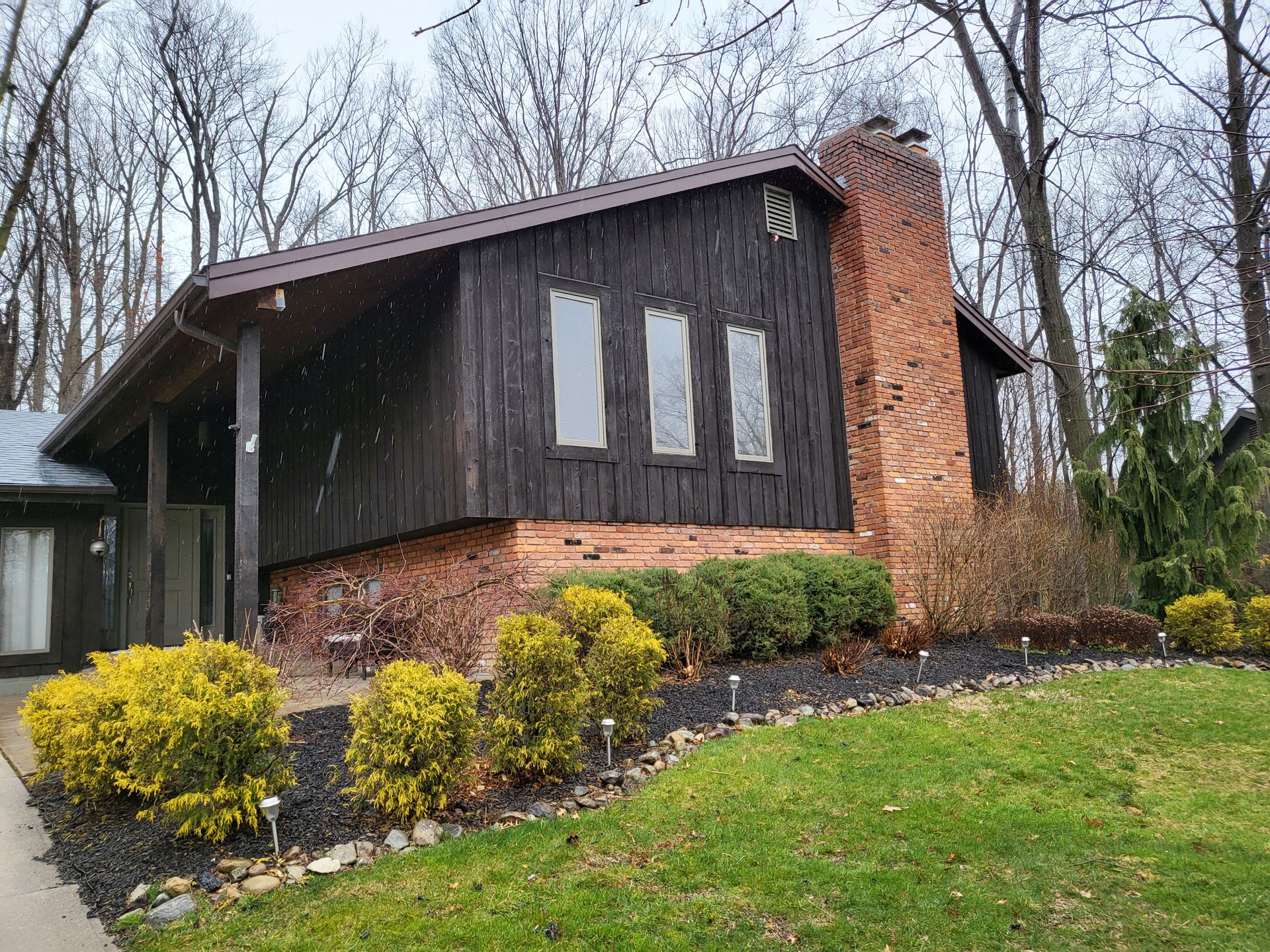 Dark brown wooden house with a brick chimney and three windows, surrounded by bushes and trees.