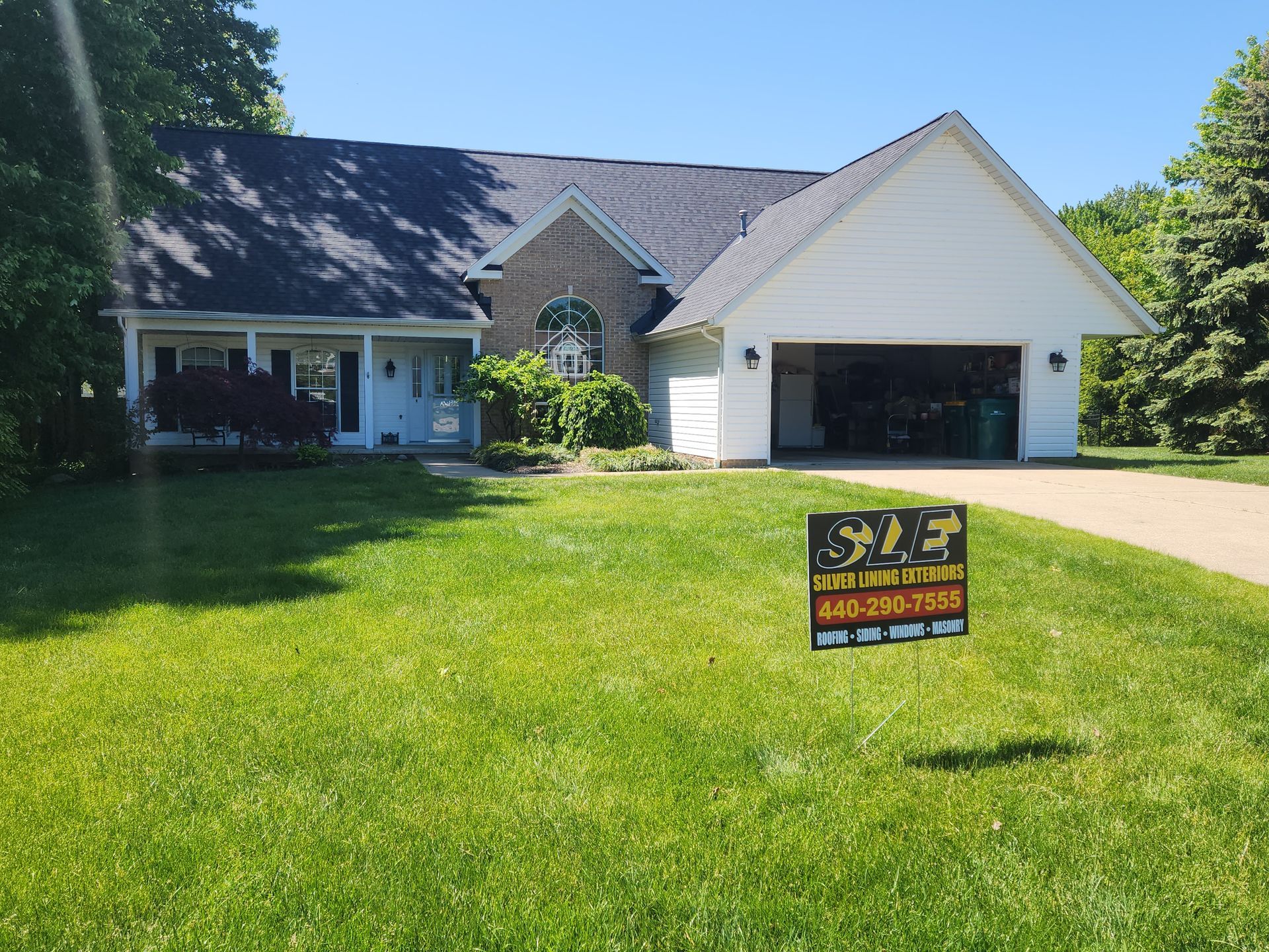 House with a dark roof, white garage, brick facade, and a sign in the yard. Sunny day.