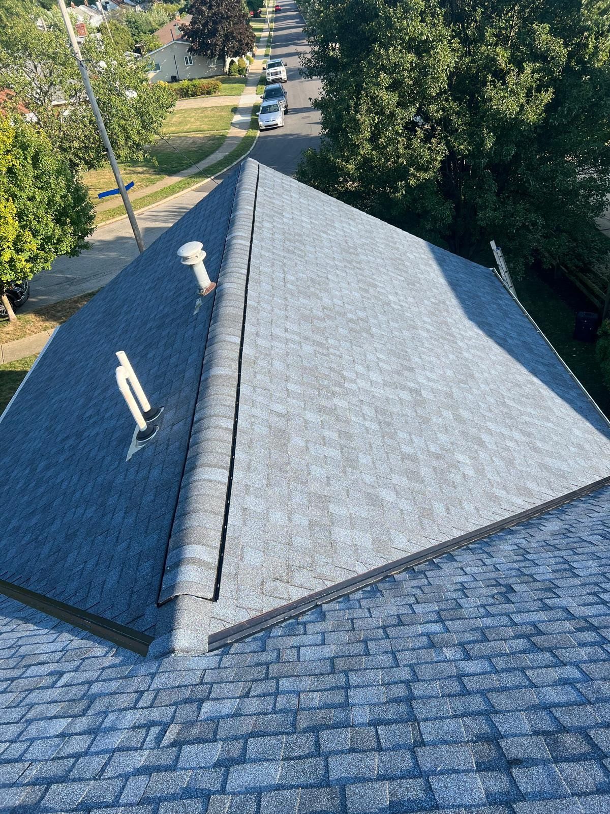 Gray asphalt shingle roof of a house, viewed from above, with a street and trees in the background.