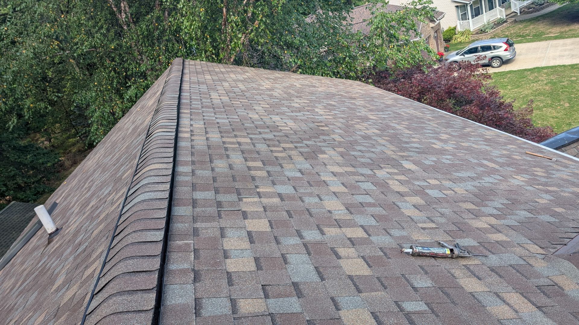 Brown shingled roof with a ridge, looking towards green trees and a house with a car.
