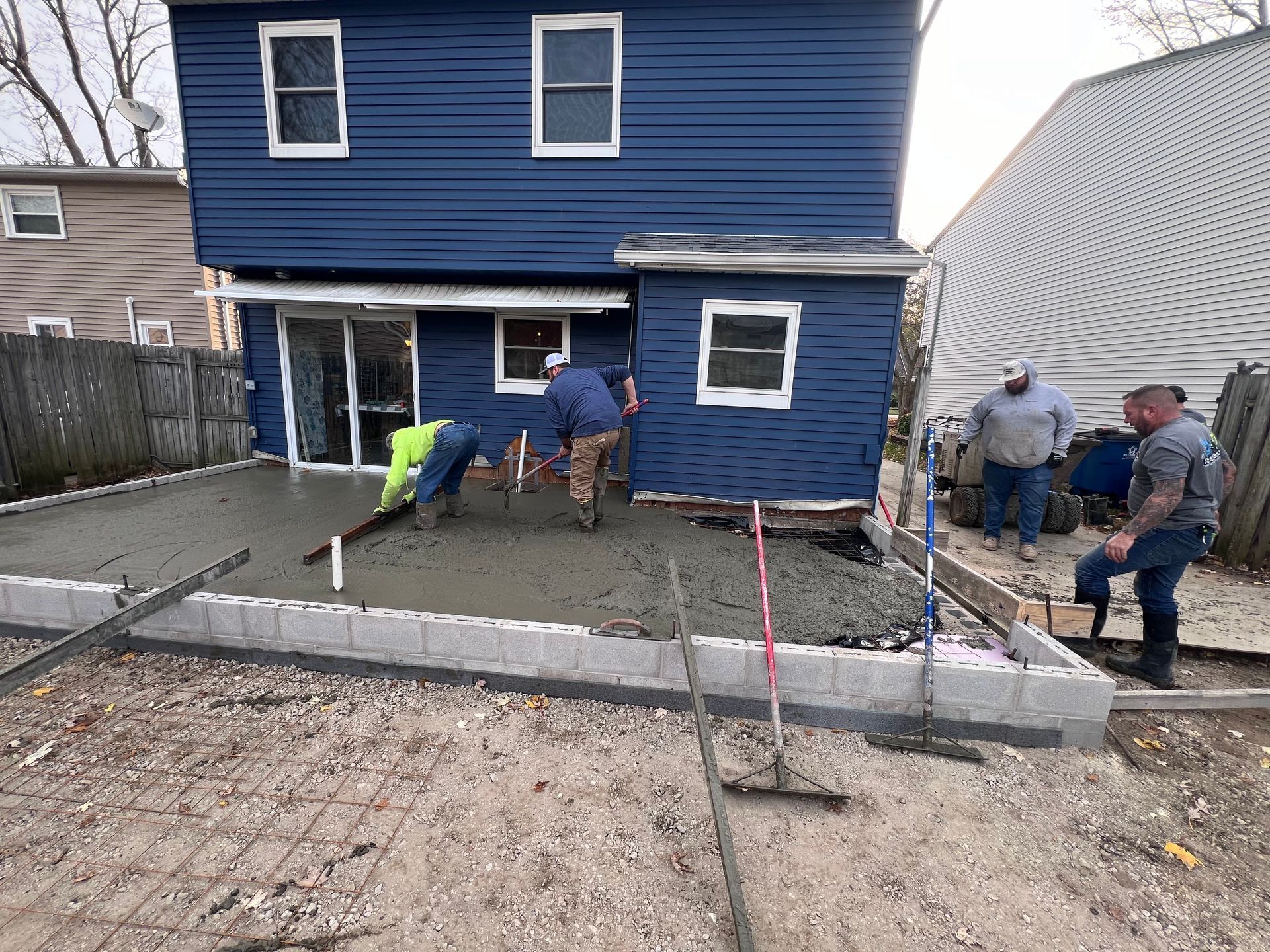 Construction workers pouring concrete patio next to a blue house; gray block border.