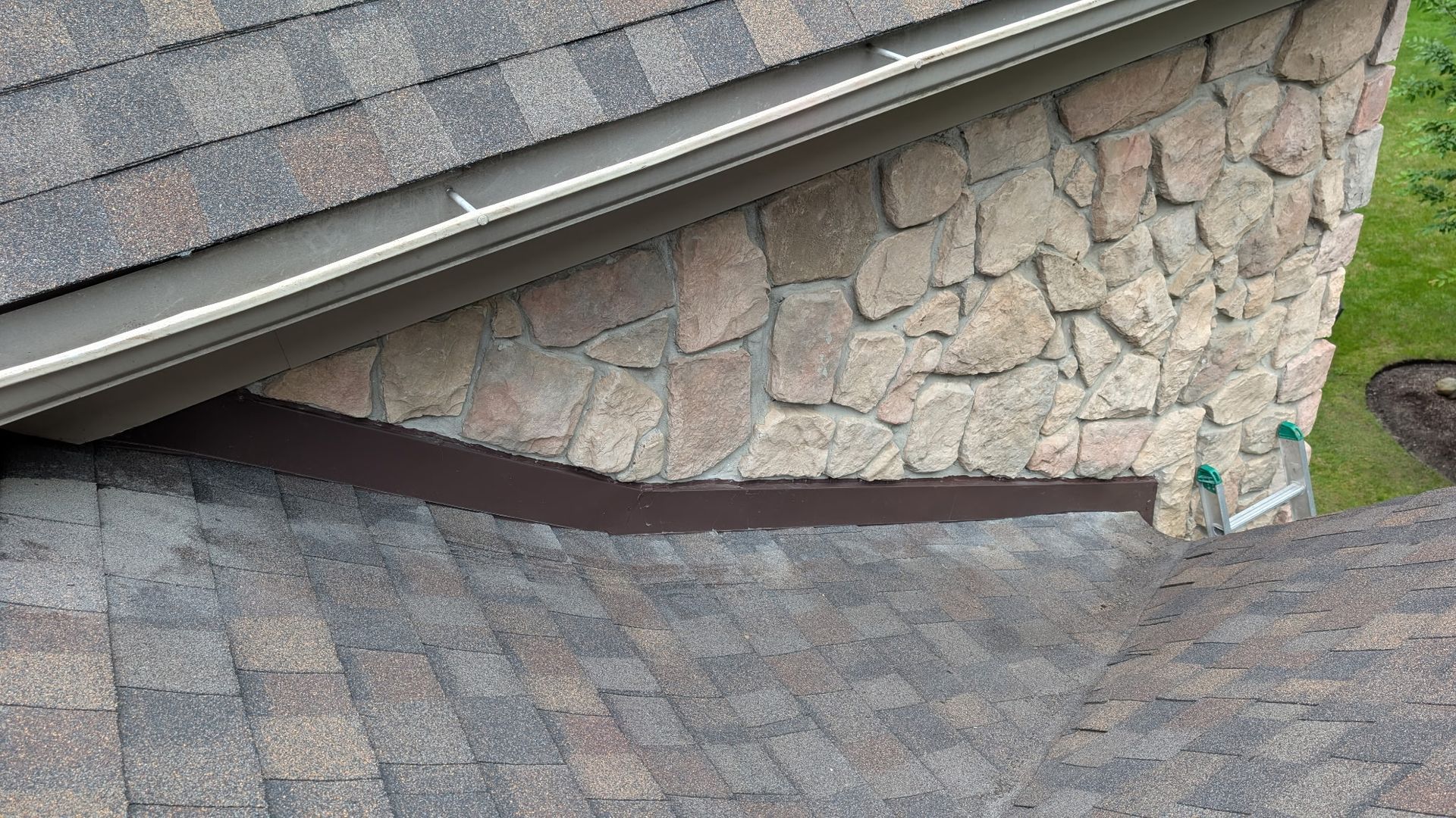 Close-up of a brown-shingled roof meeting a stone chimney. Dark brown flashing separates them.