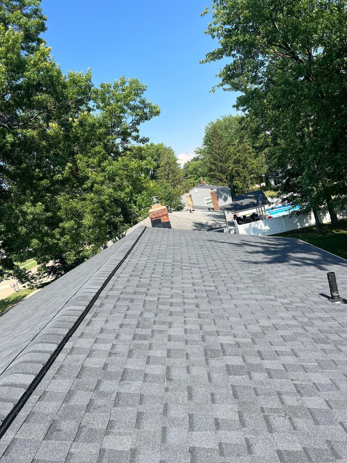 View from a roof of a gray shingled roof extending towards a residential street lined with trees under a blue sky.