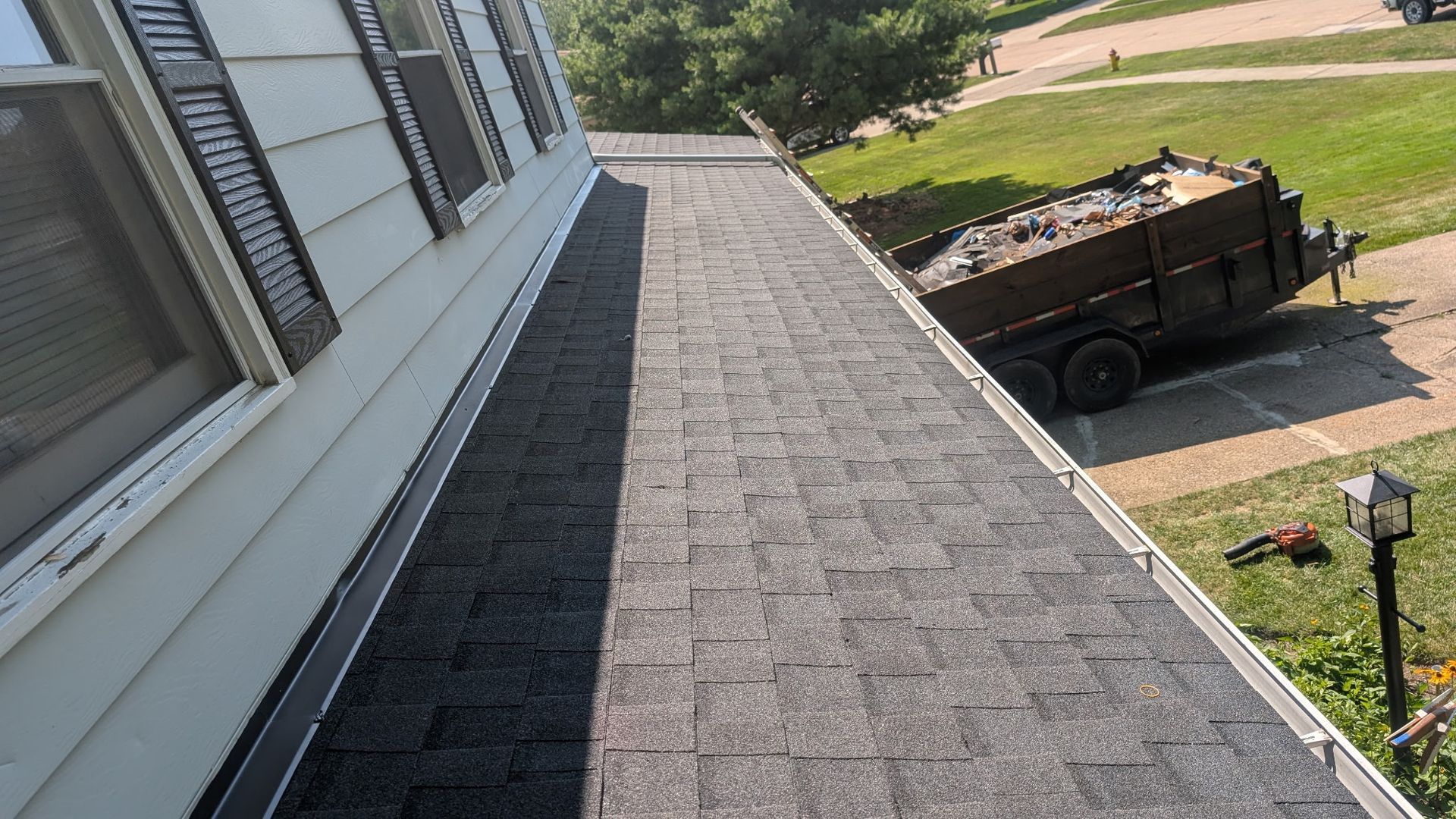Dark asphalt shingle roof on a white house. A trailer filled with debris sits next to the house.
