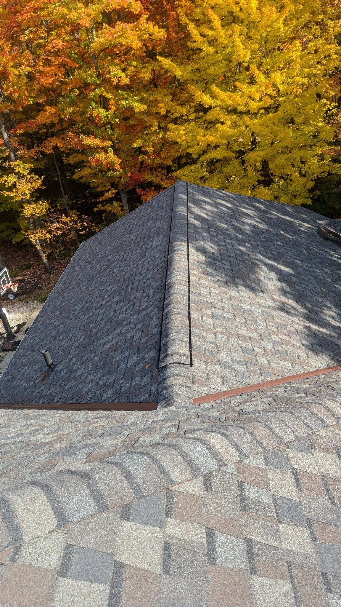 Roof with gray shingles and a ridge, against a backdrop of fall foliage.