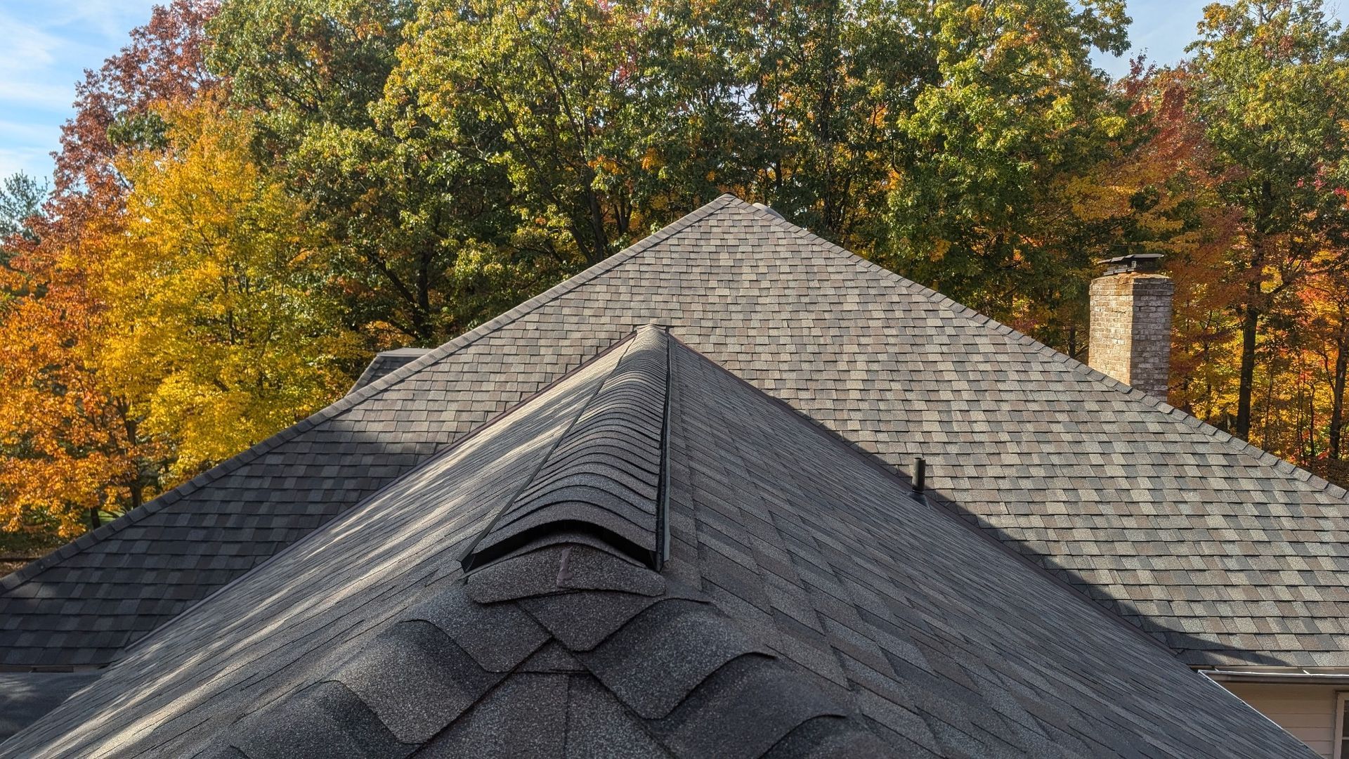 Roof of a house with gray shingles, chimney, and autumn trees in the background with colorful foliage.