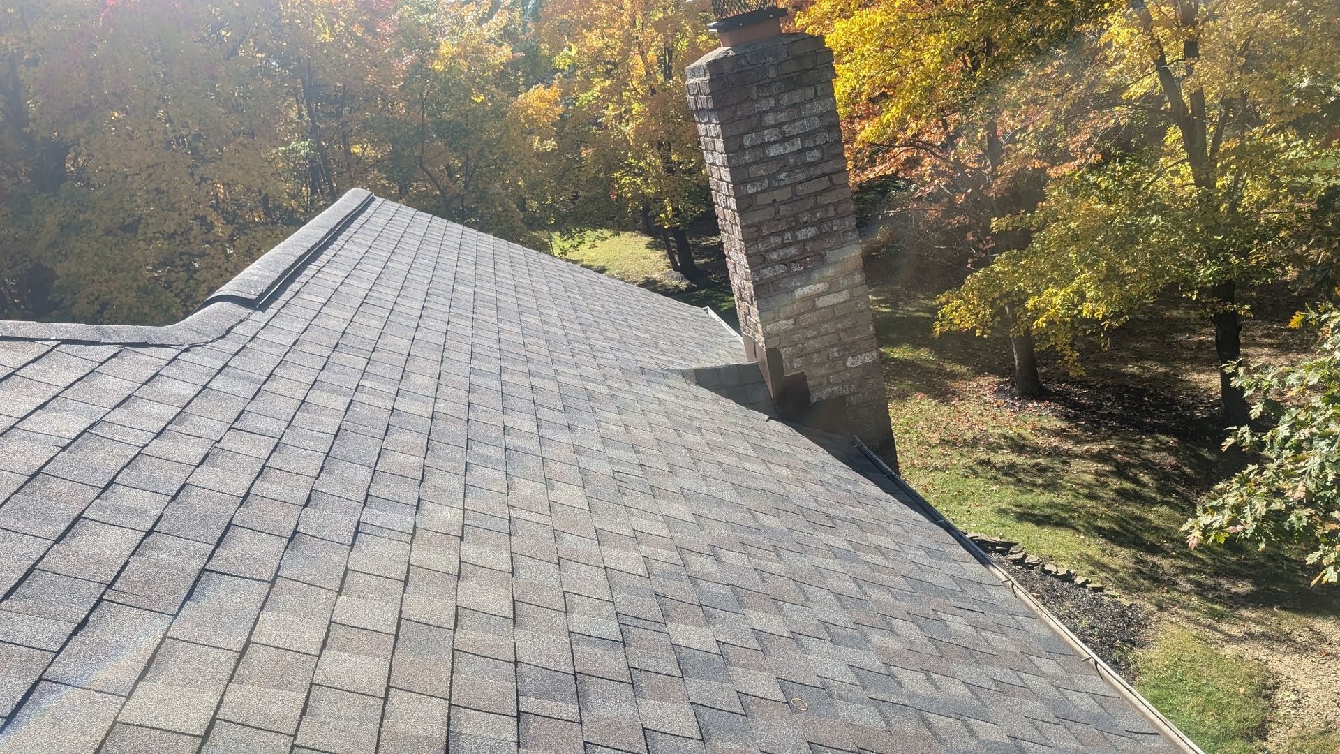 Overhead view of a roof with asphalt shingles and a brick chimney, surrounded by trees with fall foliage.