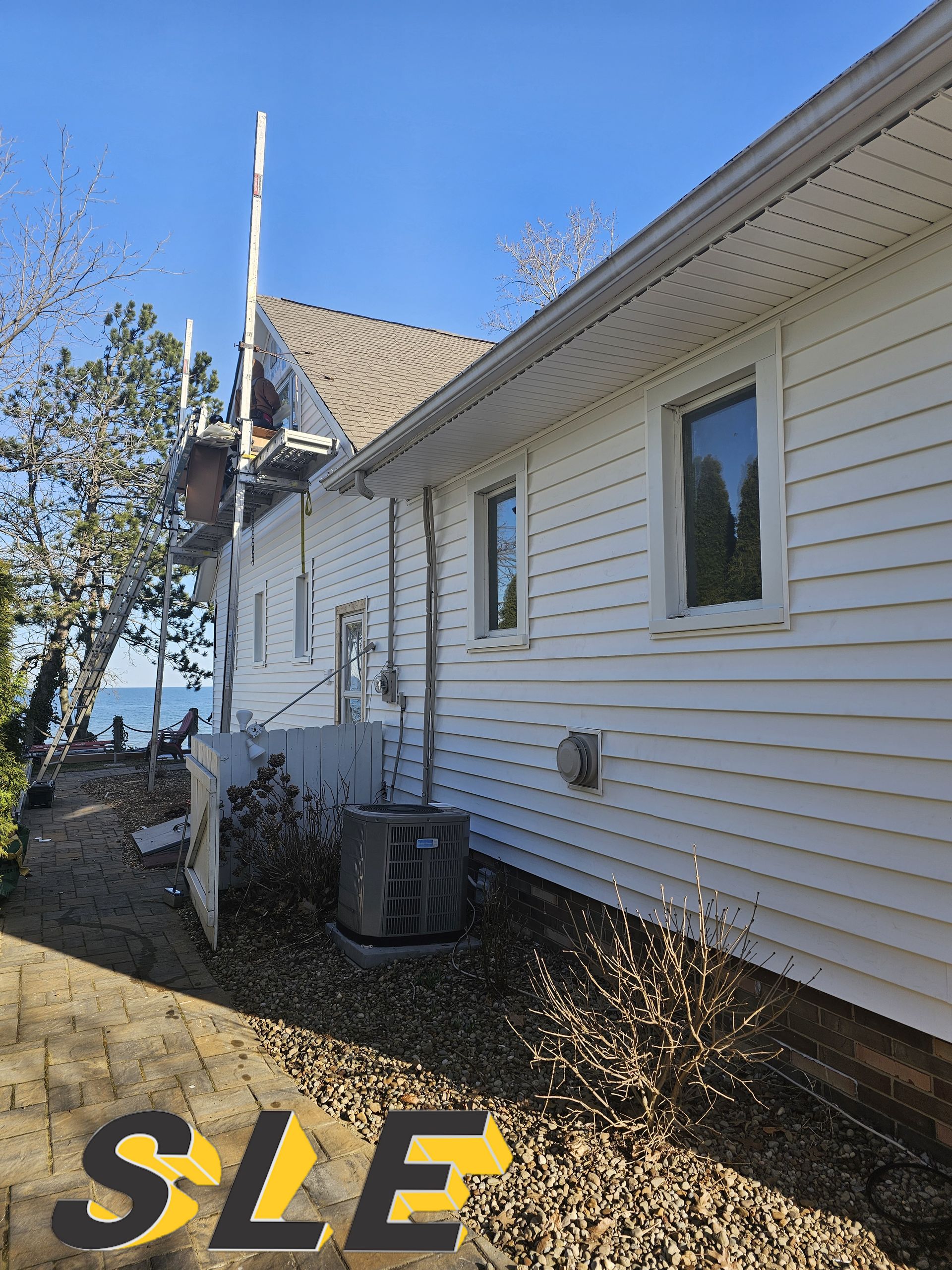 White house with siding, brown roof, by water, with an AC unit.