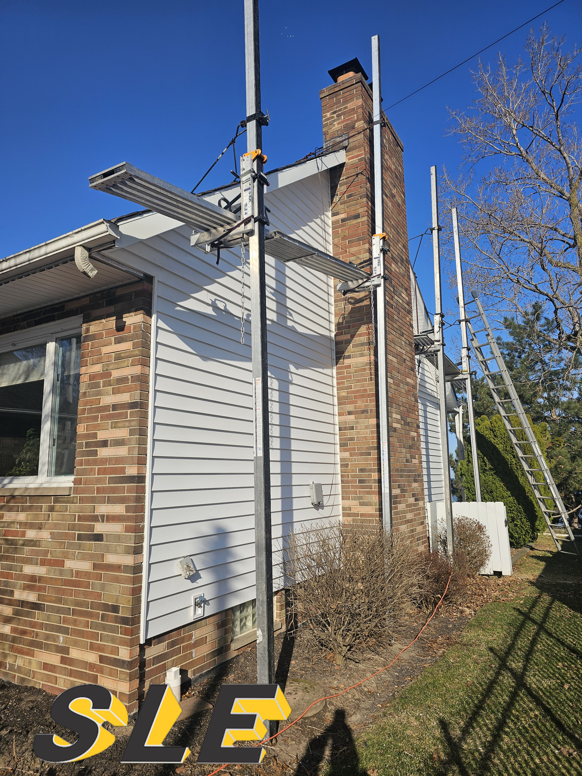 Scaffolding supports chimney repair on a brick and white-sided house under a clear blue sky.