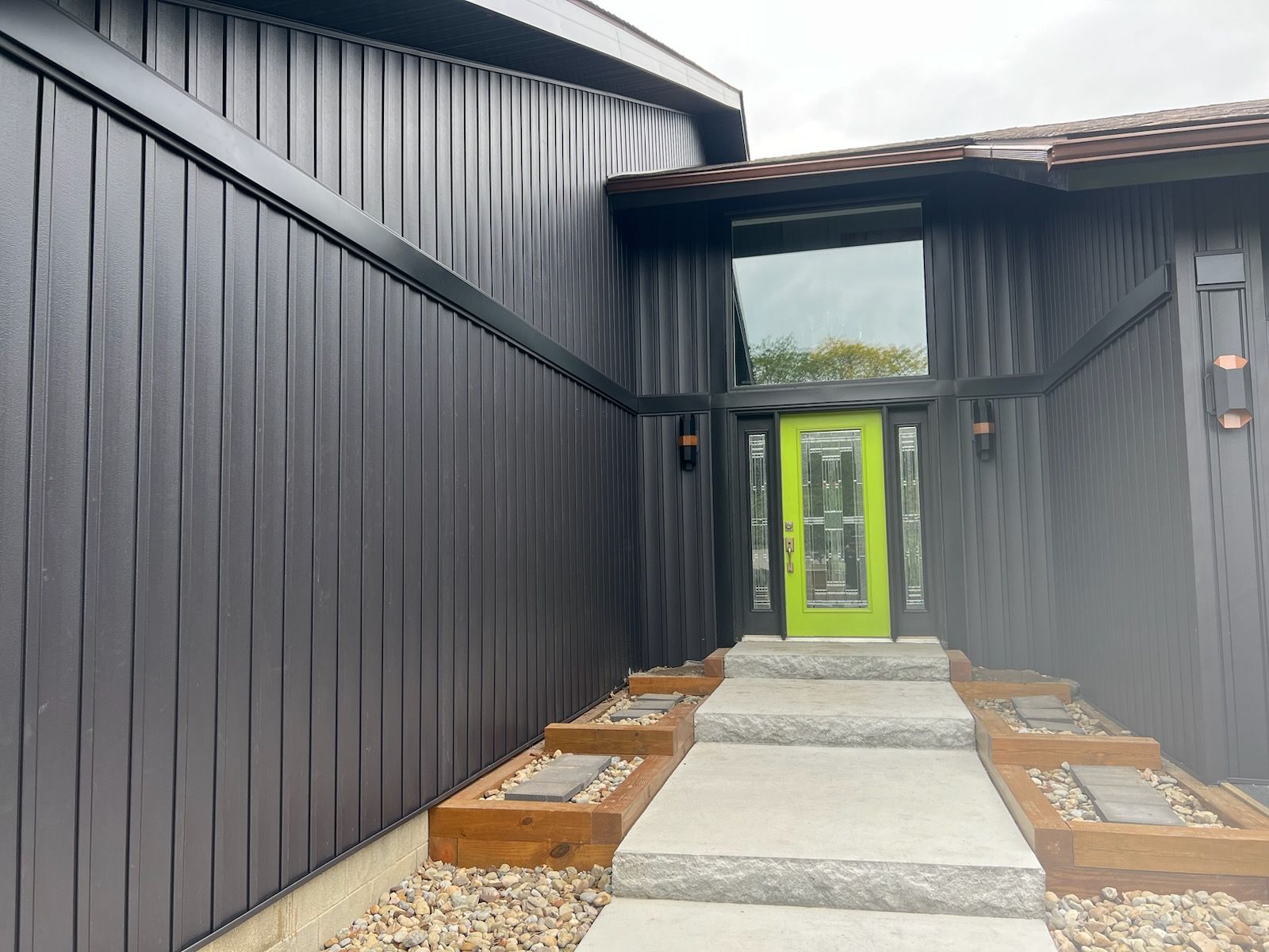 Black-sided house with a lime-green front door. Grey steps and a window lead to the entrance.