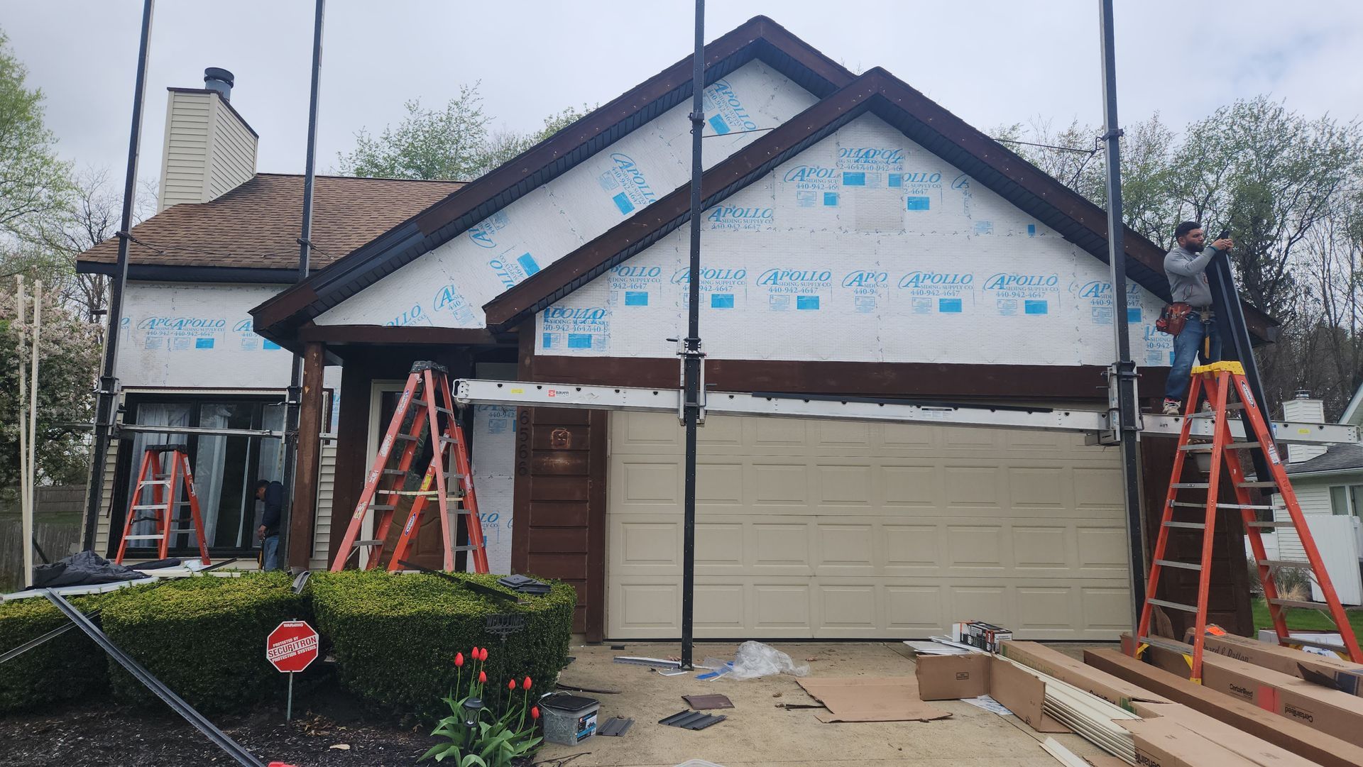 House exterior under renovation; brown siding, garage door, and roof; workers on ladders.