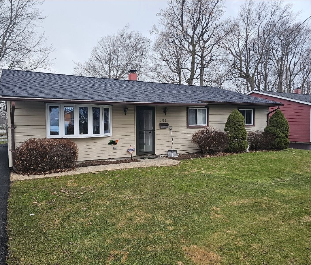 Ranch-style house with tan siding, a dark roof, and a green lawn. Bushes and trees surround the house.