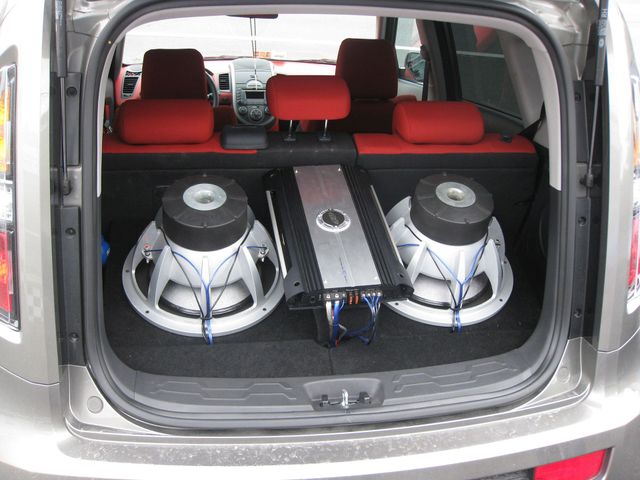 Car trunk with two large subwoofers and an amplifier installed on a carpeted floor, red seats visible inside