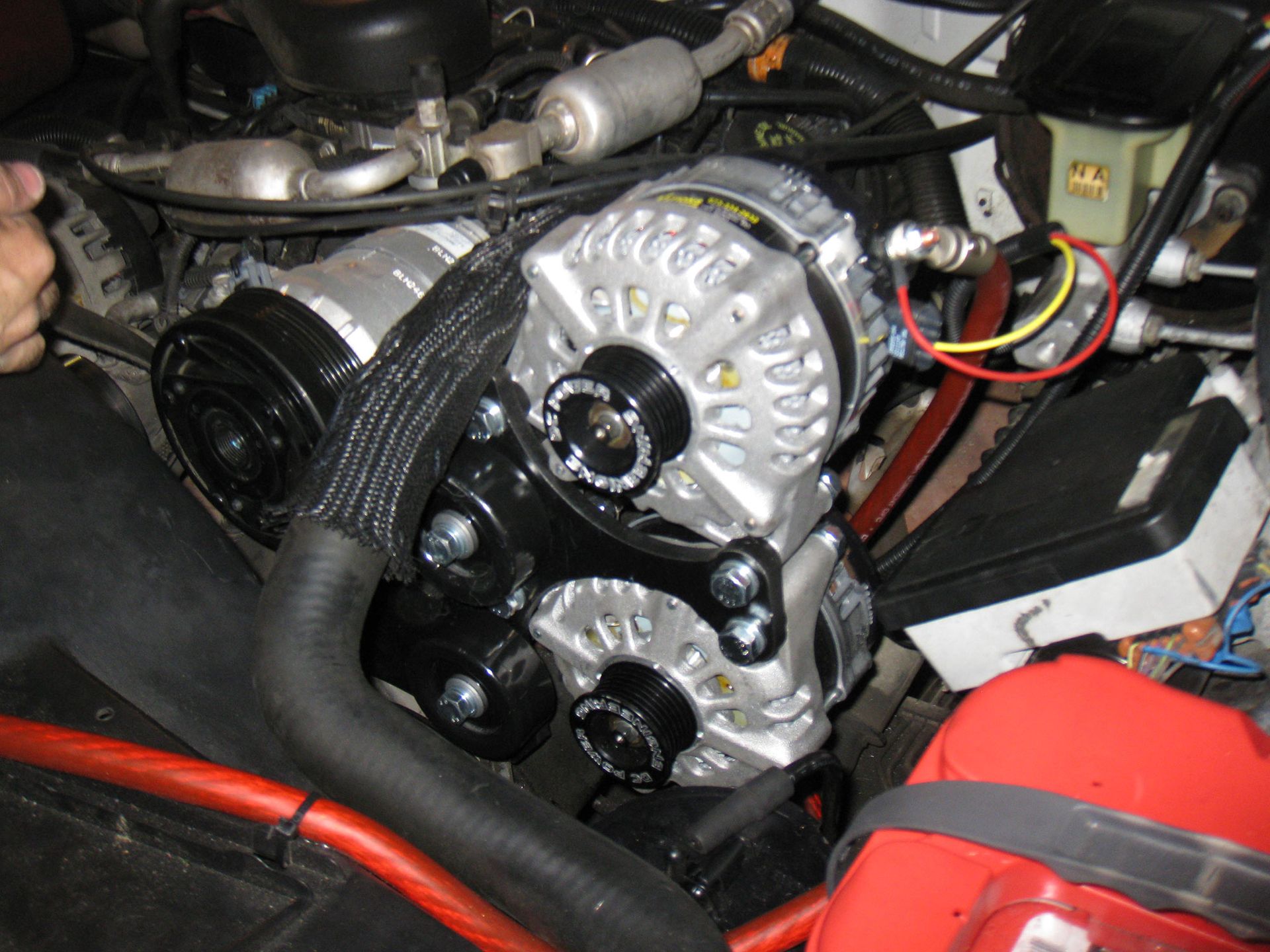 Close-up of a car engine with a silver alternator, black hoses, and red wiring in the engine bay