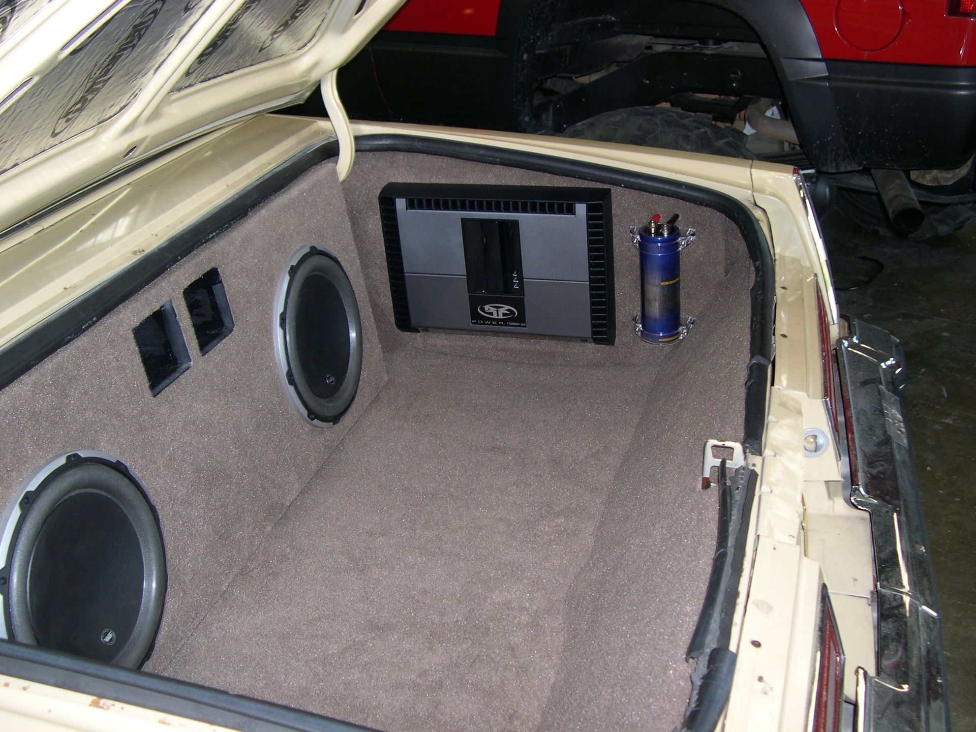 Car trunk audio setup with beige carpet, two large speakers, and an amplifier panel