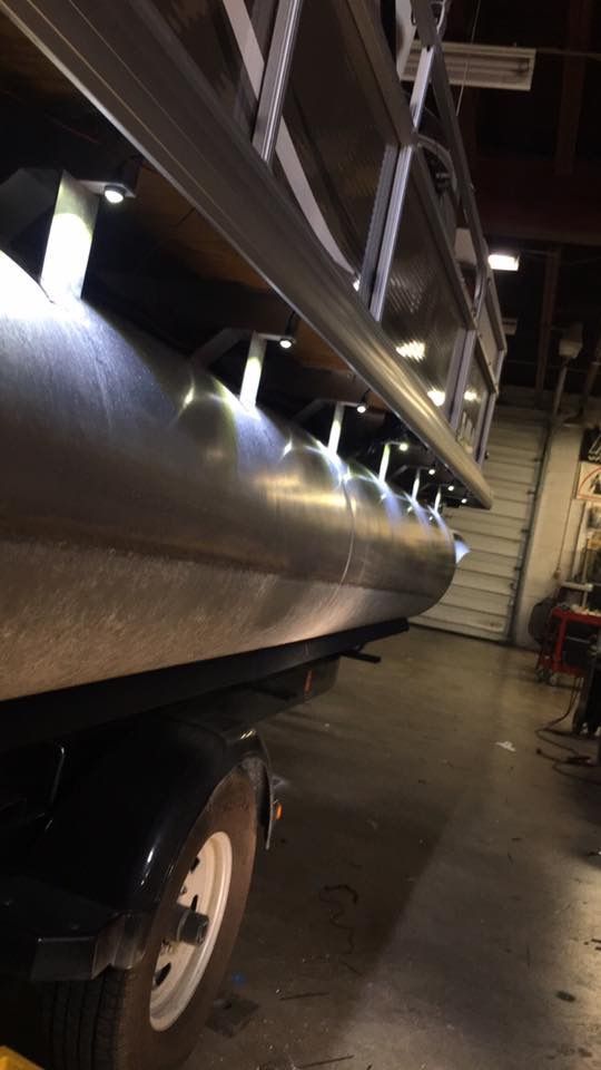 A silver pontoon boat stored indoors in a dim garage, with stairs and lights visible in the background.