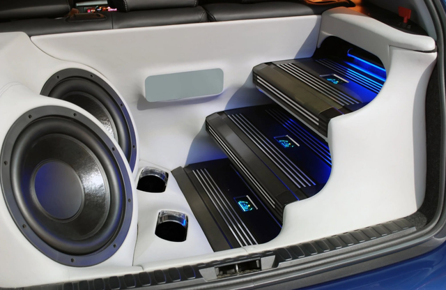 Custom car audio system with large subwoofer, black speaker pods, and blue-lit steps in a trunk compartment