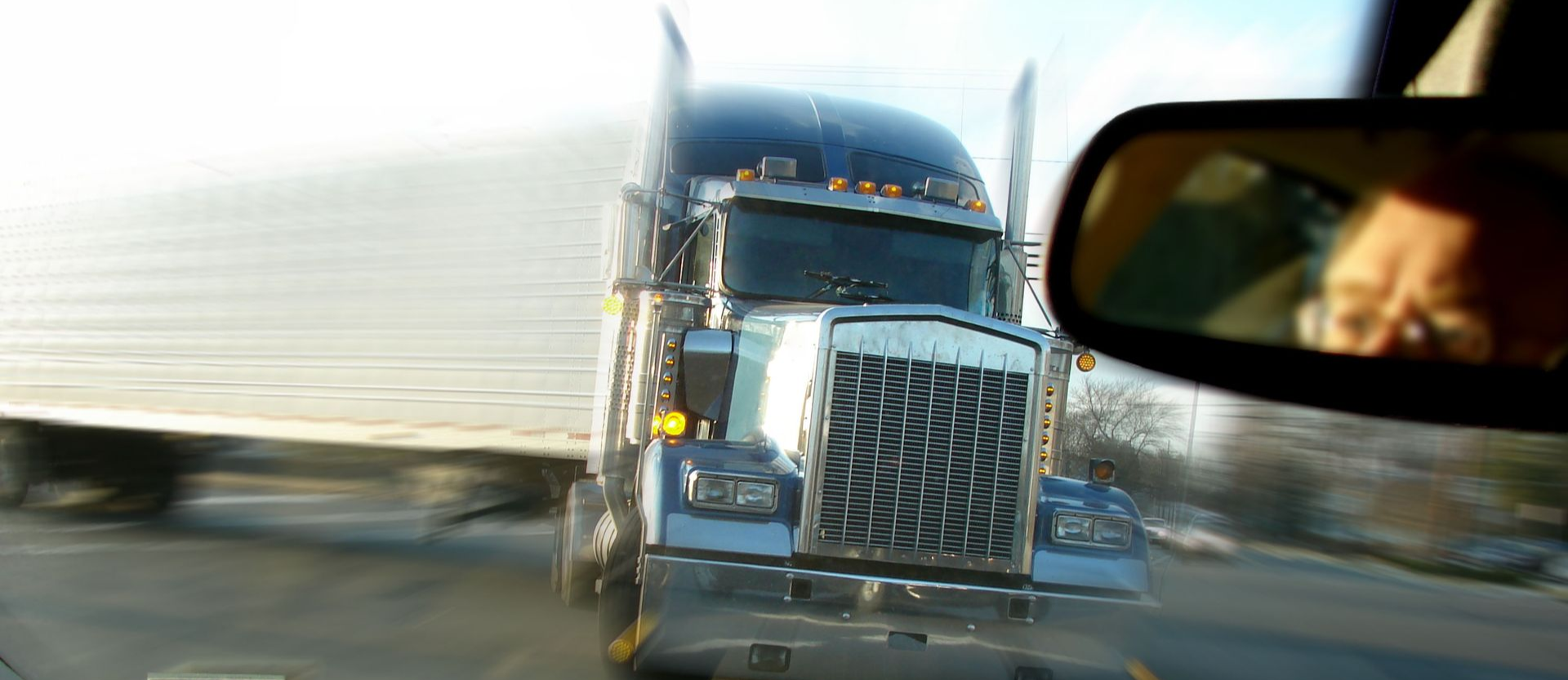 Blue semi-truck driving on a highway, seen from a car with a blurred side mirror in the foreground