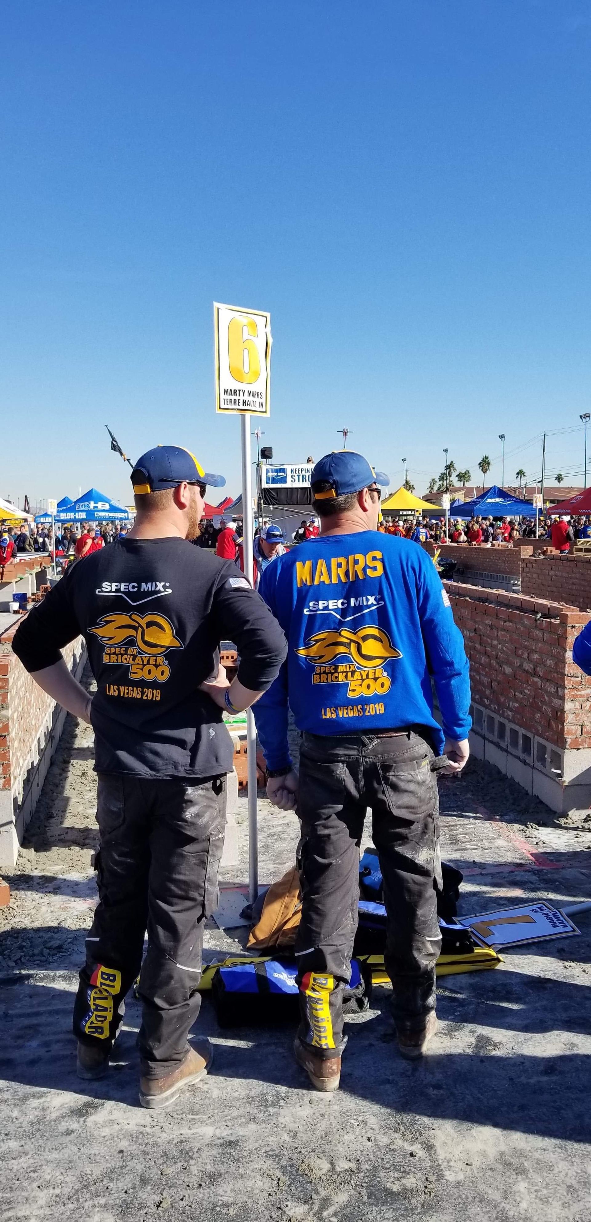Two men in blue shirts are standing next to each other on a dirt road.