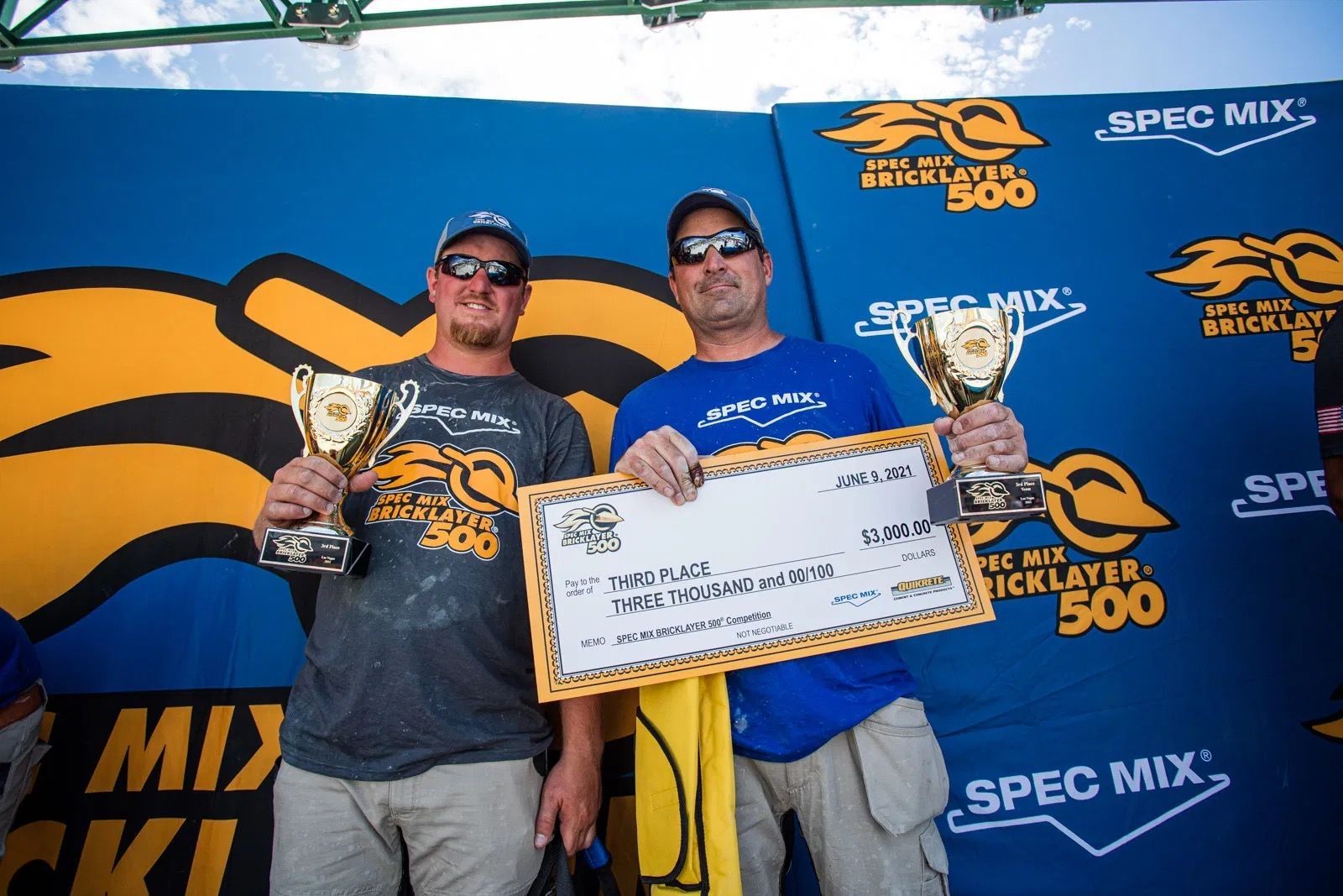 Two men holding trophies and a check in front of a spec mix sign