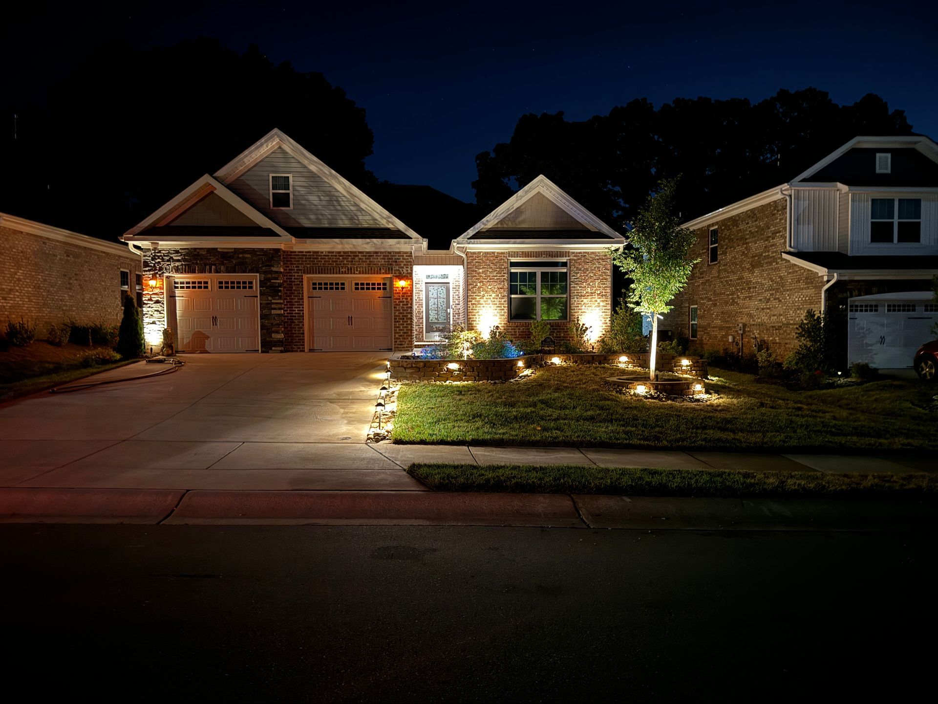 A house at night with landscape lighting, illuminating the front yard, bushes, and garage doors.