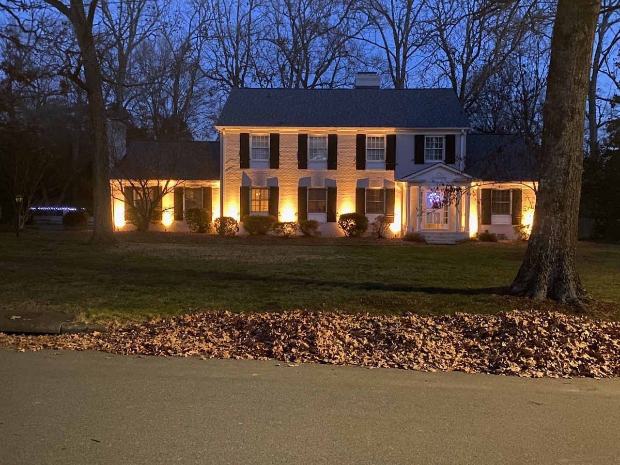 A two-story house illuminated with warm lights, framed by bare trees, and fallen leaves in the foreground.