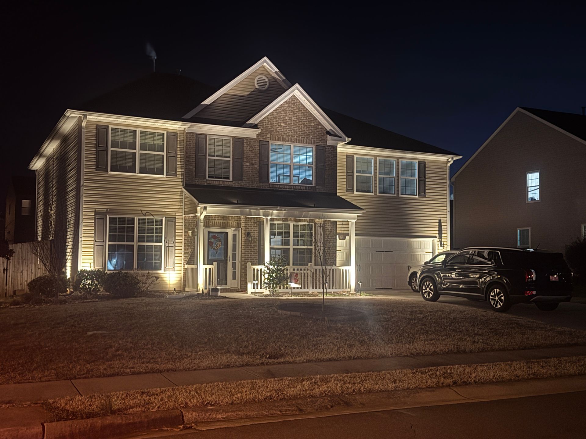 A two-story house at night with lights illuminating its facade and a vehicle parked in the driveway.