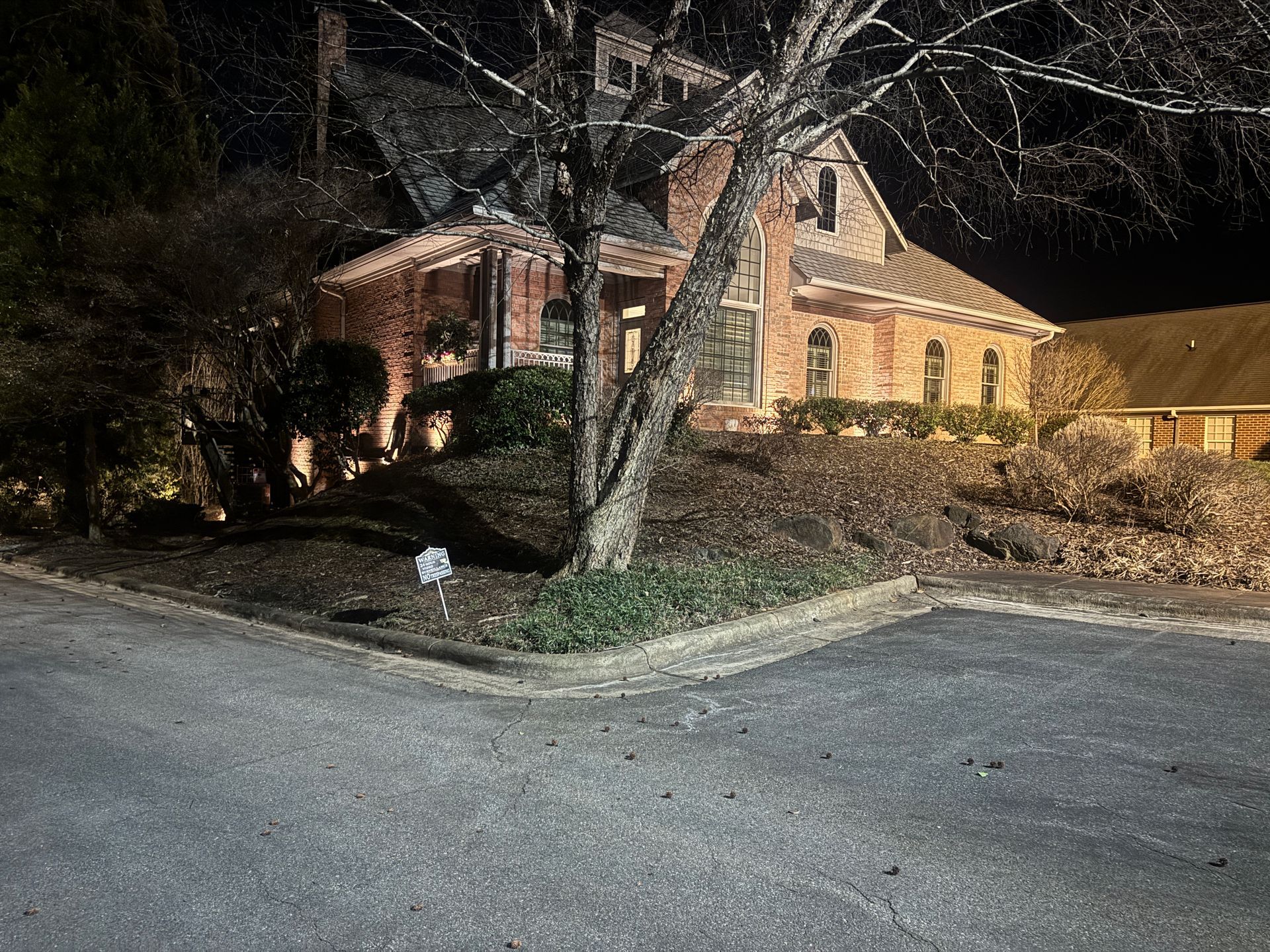 Night shot of a brick house with a tree in front. The house has multiple windows, and is lit. A street is in front.