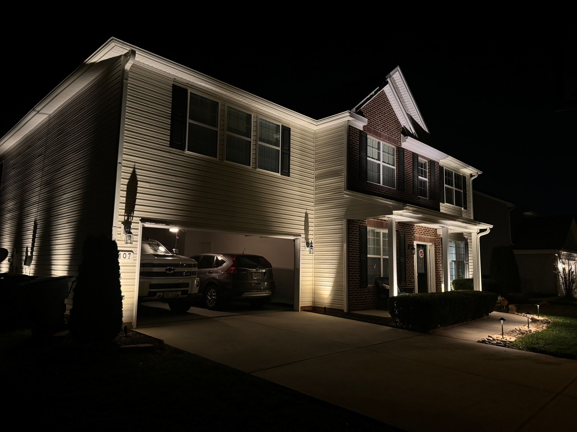 Night view of a two-story house with outdoor lighting illuminating the facade and a garage with cars inside.