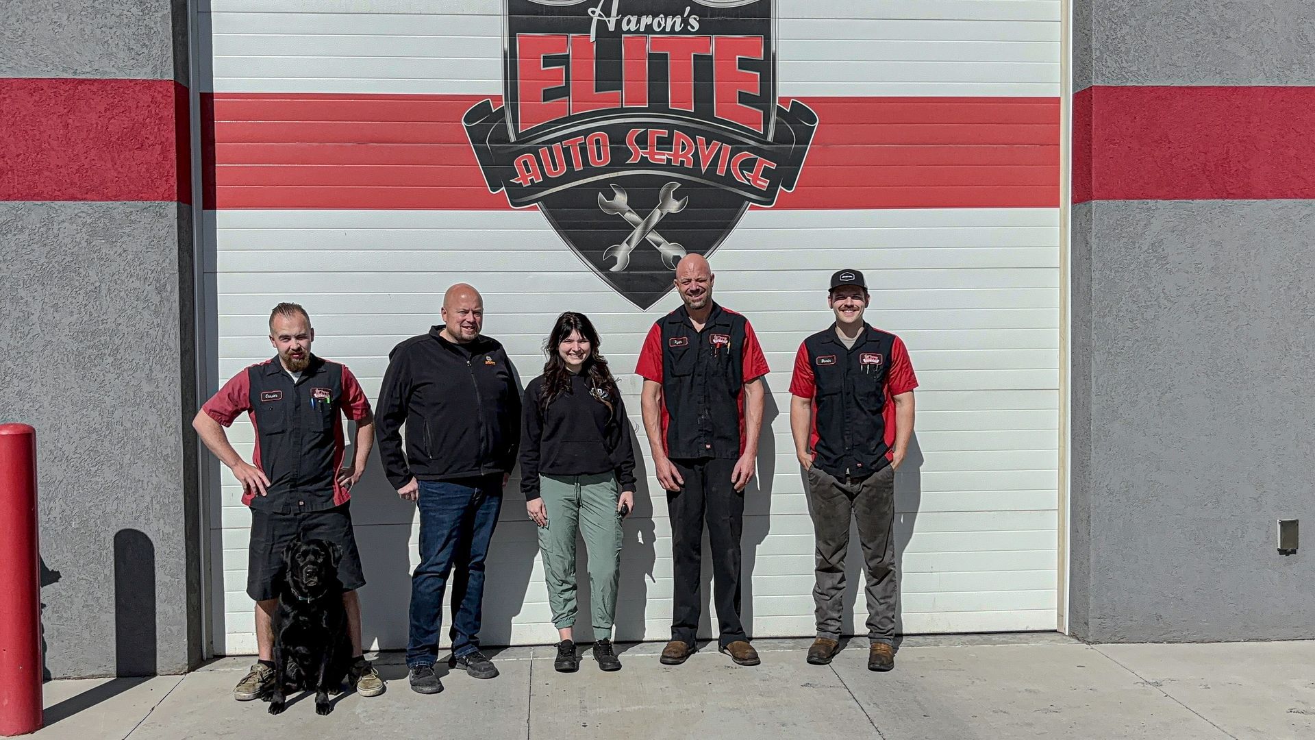 A group of five people in auto shop uniforms standing in front of a garage door with their business logo.