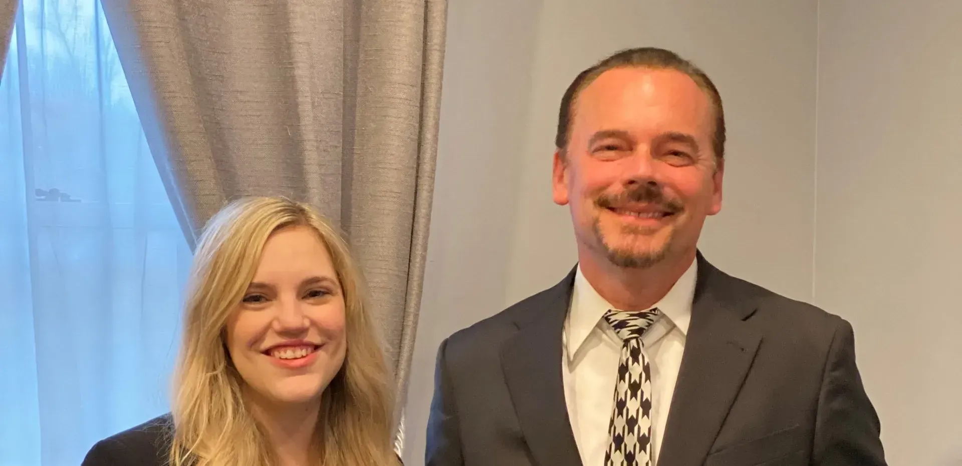A smiling blonde woman and a smiling man in a suit stand side-by-side.
