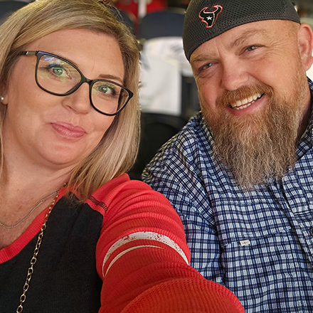 A man and a woman are taking a selfie together . the man is wearing a hat with a bull on it.