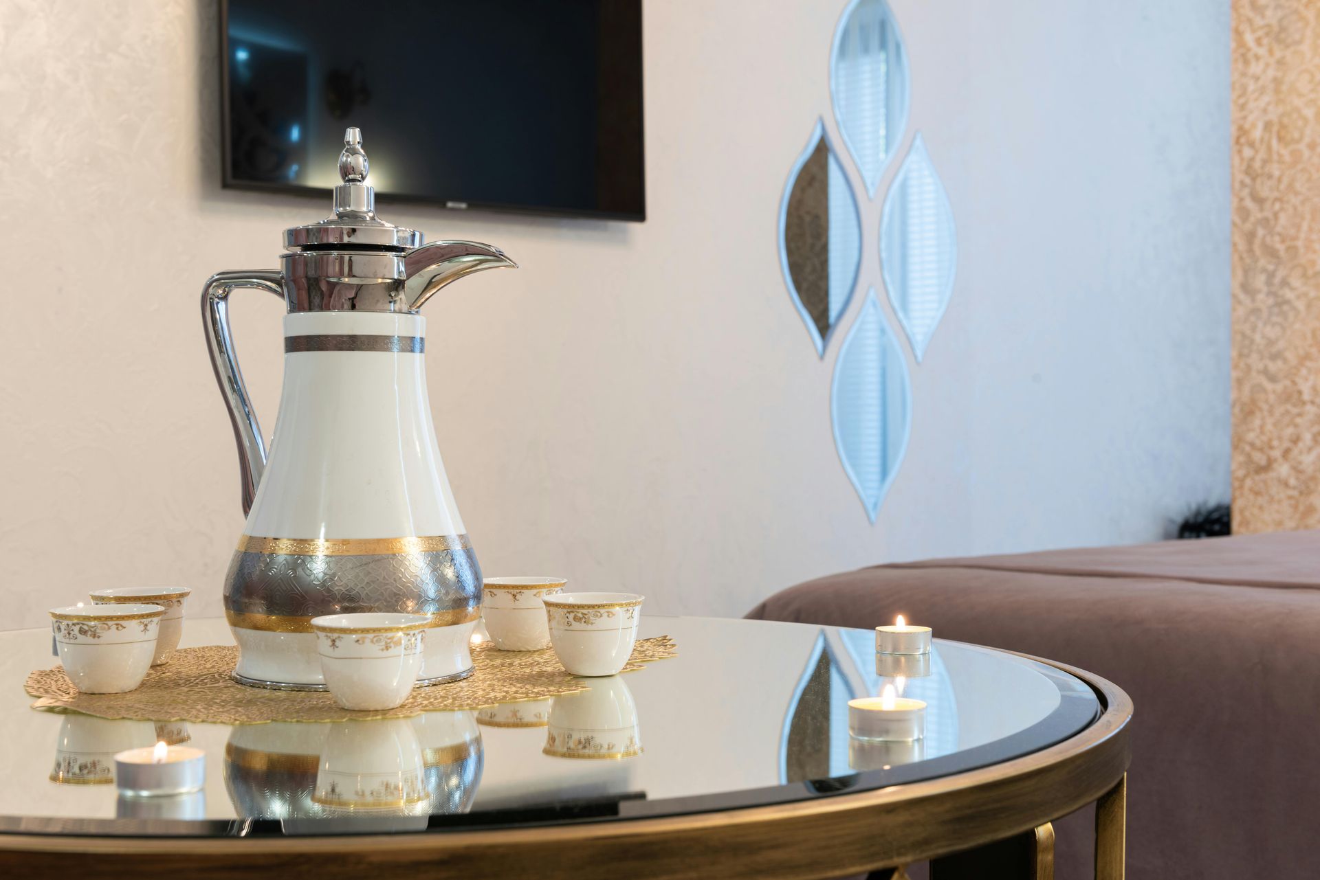 A white and gold coffee carafe with small matching cups and tea lights on a glass table in a living room.