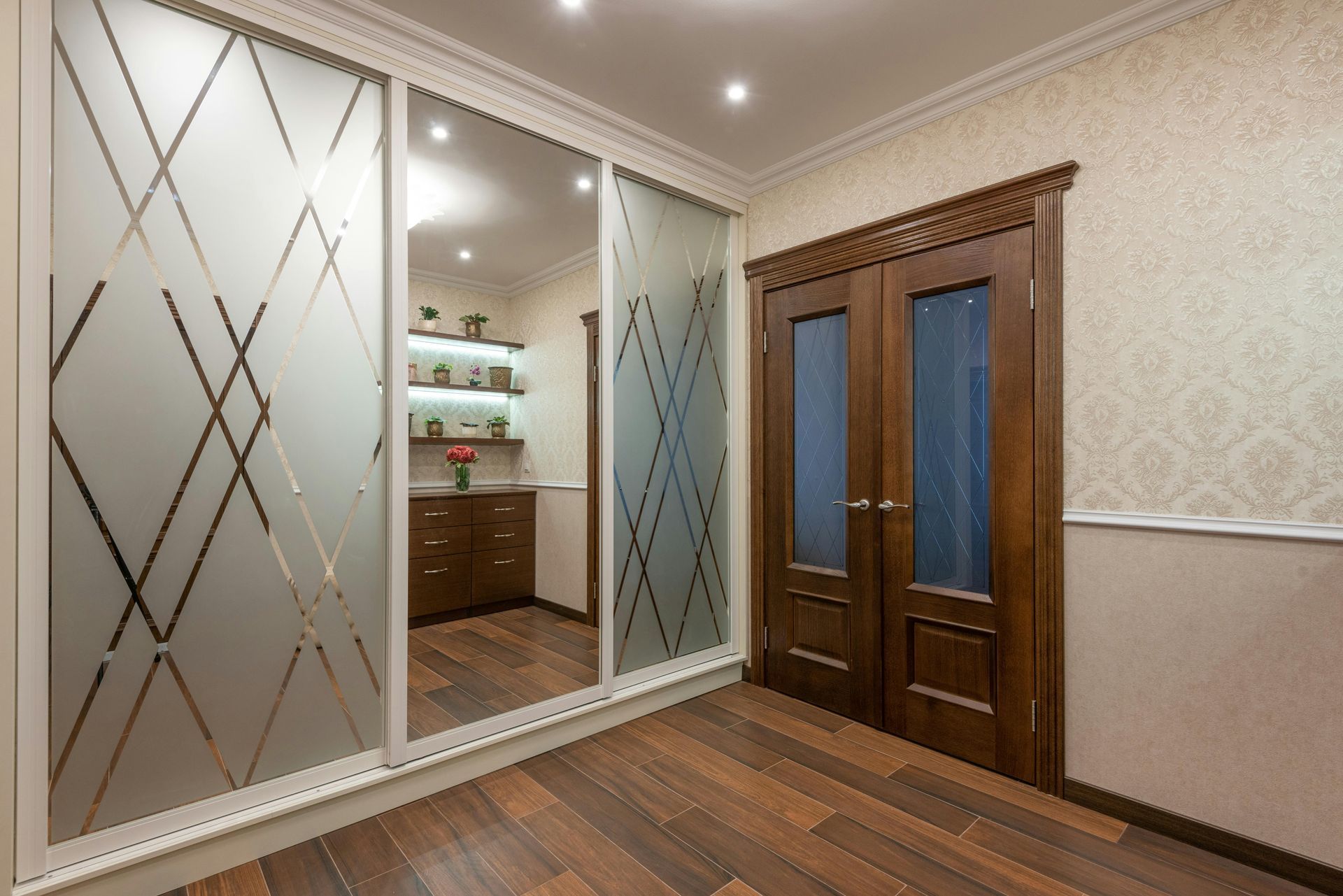 A hallway features a large sliding wardrobe with frosted geometric panels and a central mirror next to wooden double doors.