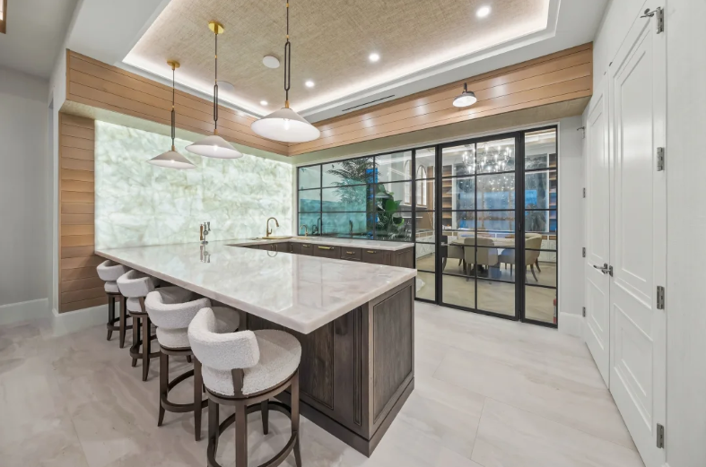 Modern kitchen with marble island, white stools, wood accents, and large glass doors opening to a patio