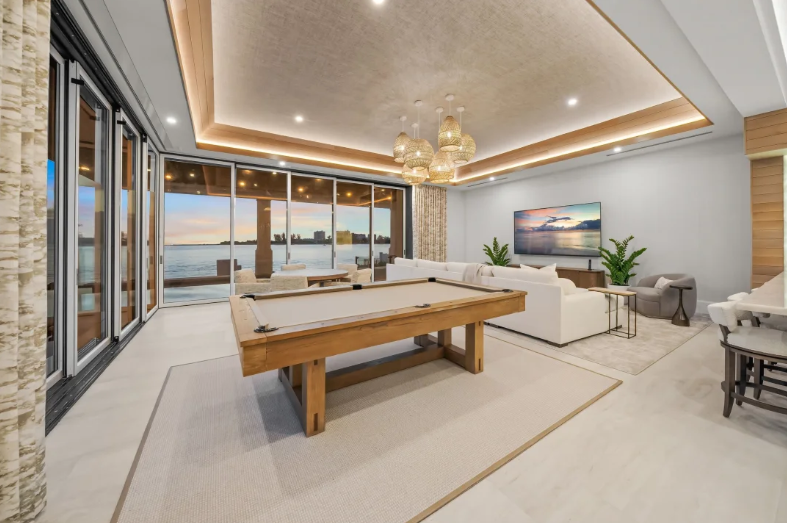 Modern living room with pool table, sofa, TV, and large windows overlooking the water