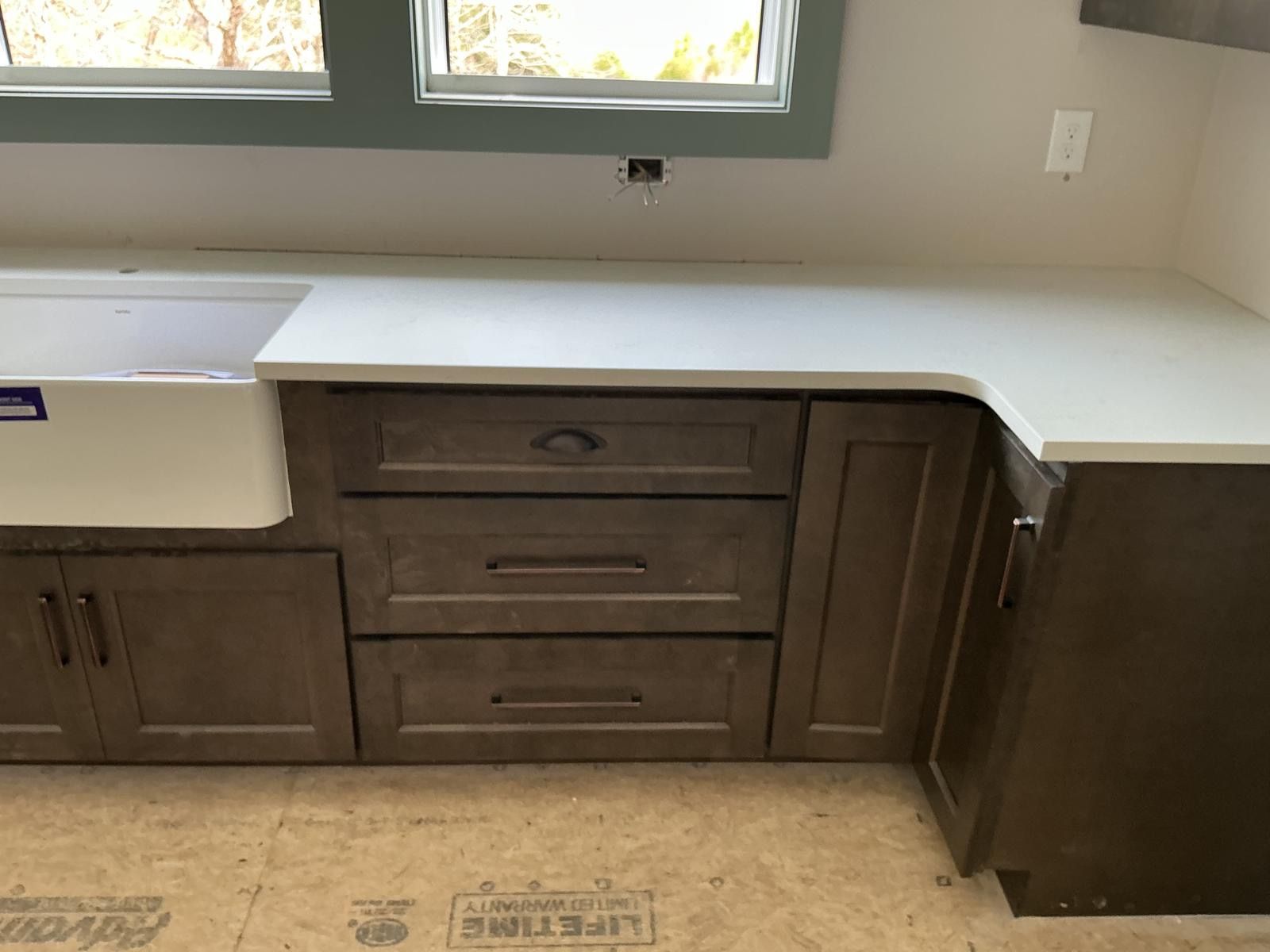 Kitchen base cabinets and white countertop under a window, with unfinished flooring and a sink on the left.