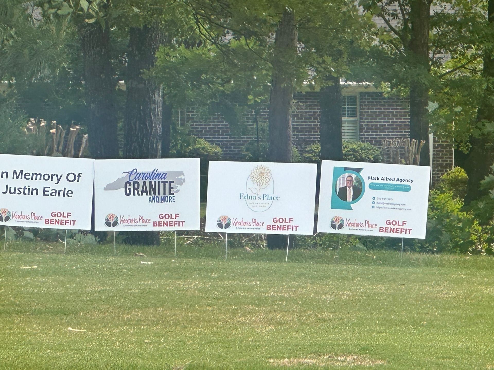 Memorial sponsor banners on a fence in a grassy park, with trees and a building behind.